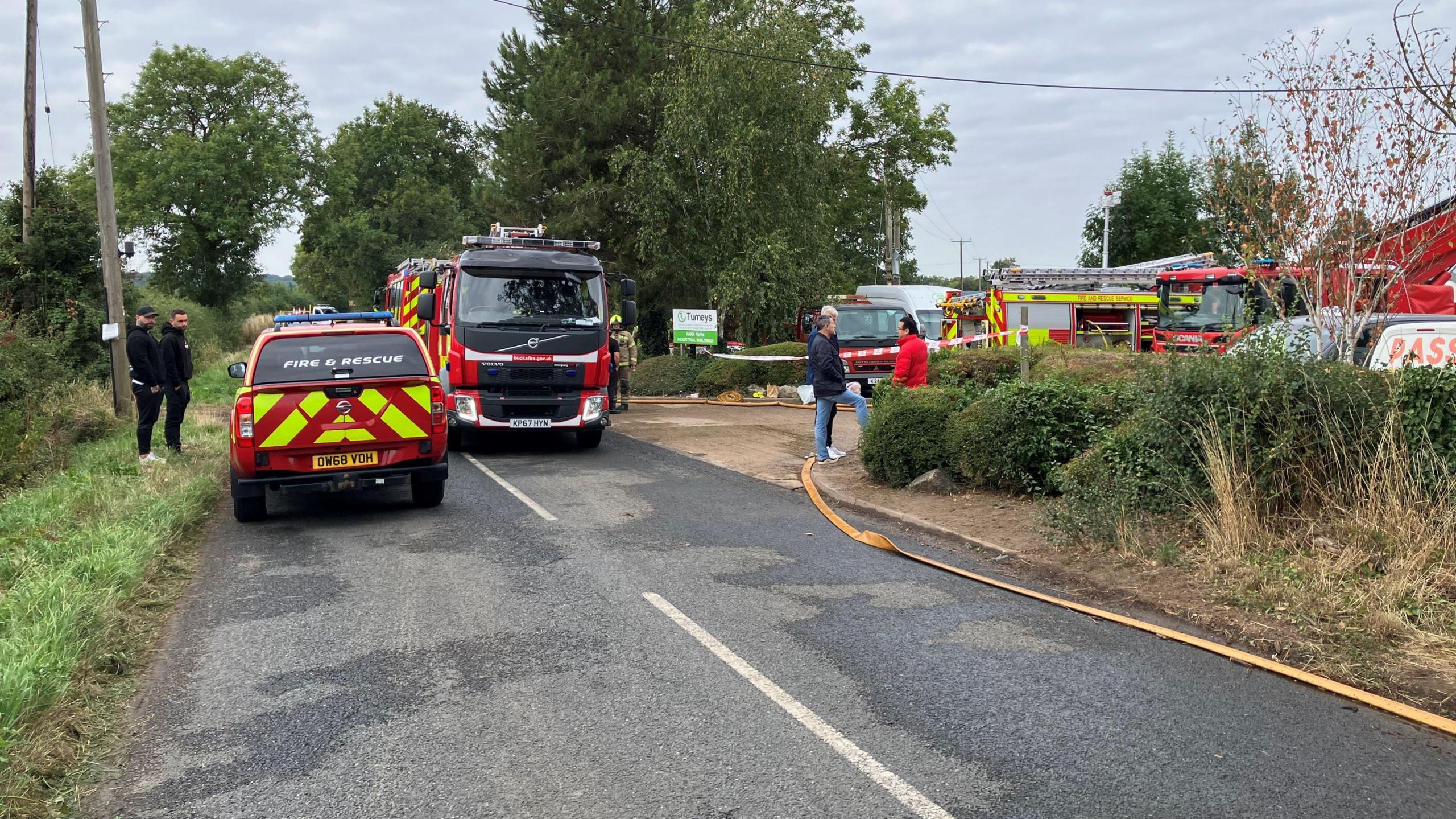Two fire engines and a car parked on the road. There are people standing outside the vehicles.