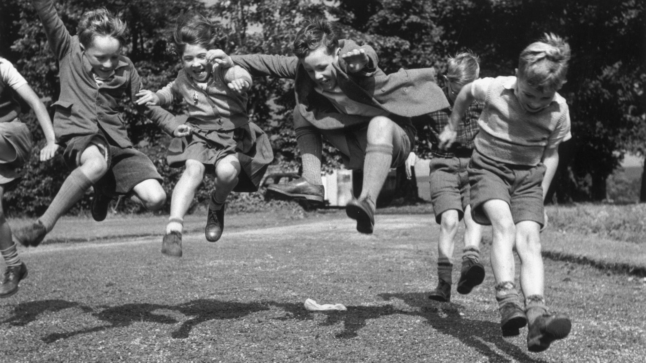 A black and white image of young children linking hands as they jump and play. They are a mixture of boys and girls and are wearing shorts or skirts. The photograph is taken in a garden which is bordered with a high hedge
