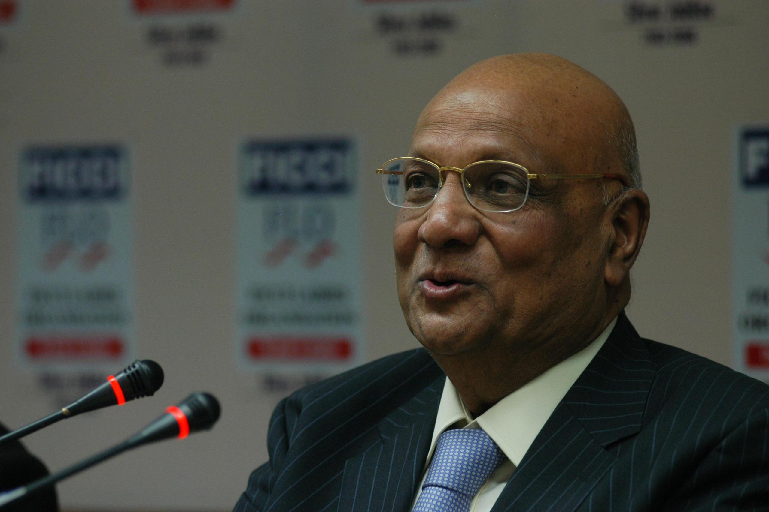 A close-up of a man wearing glasses, a white shirt, a blue tie and a pin-striped suit. He sits in front of two small microphones.