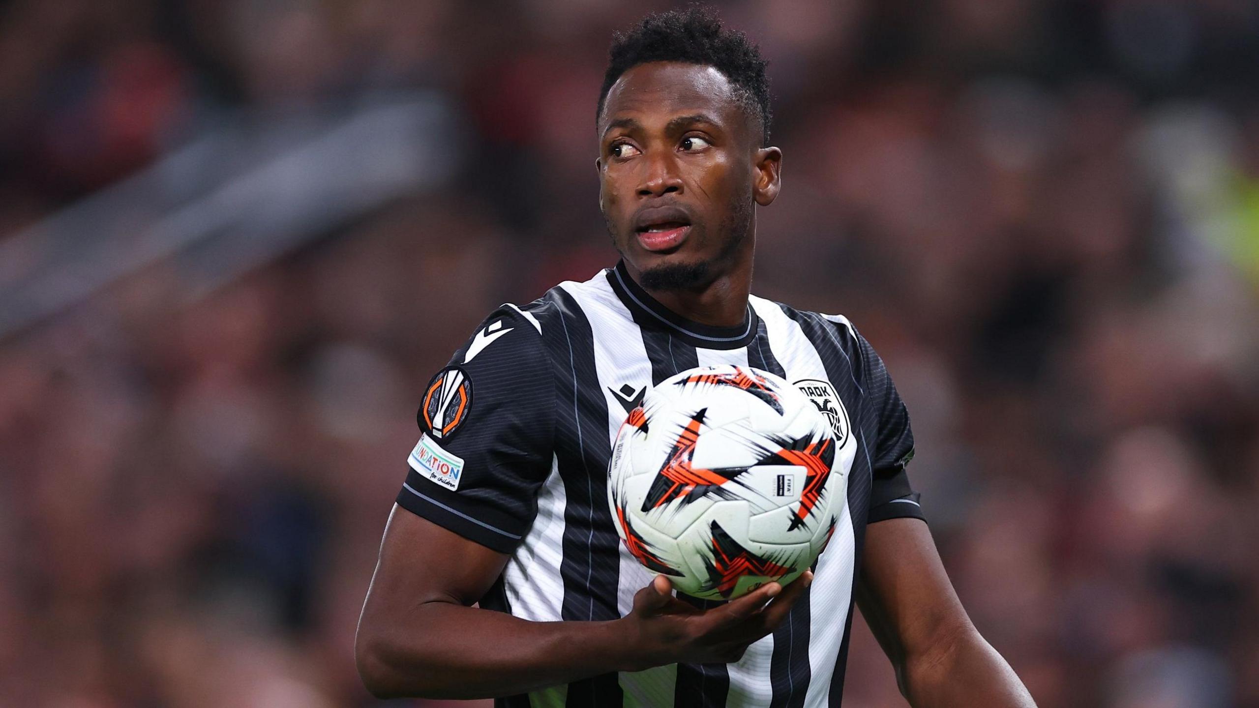 Baba Rahman, wearing a black and white striped PAOK shirt, holds a football in the palm of his right hand as he turns his head to his right over