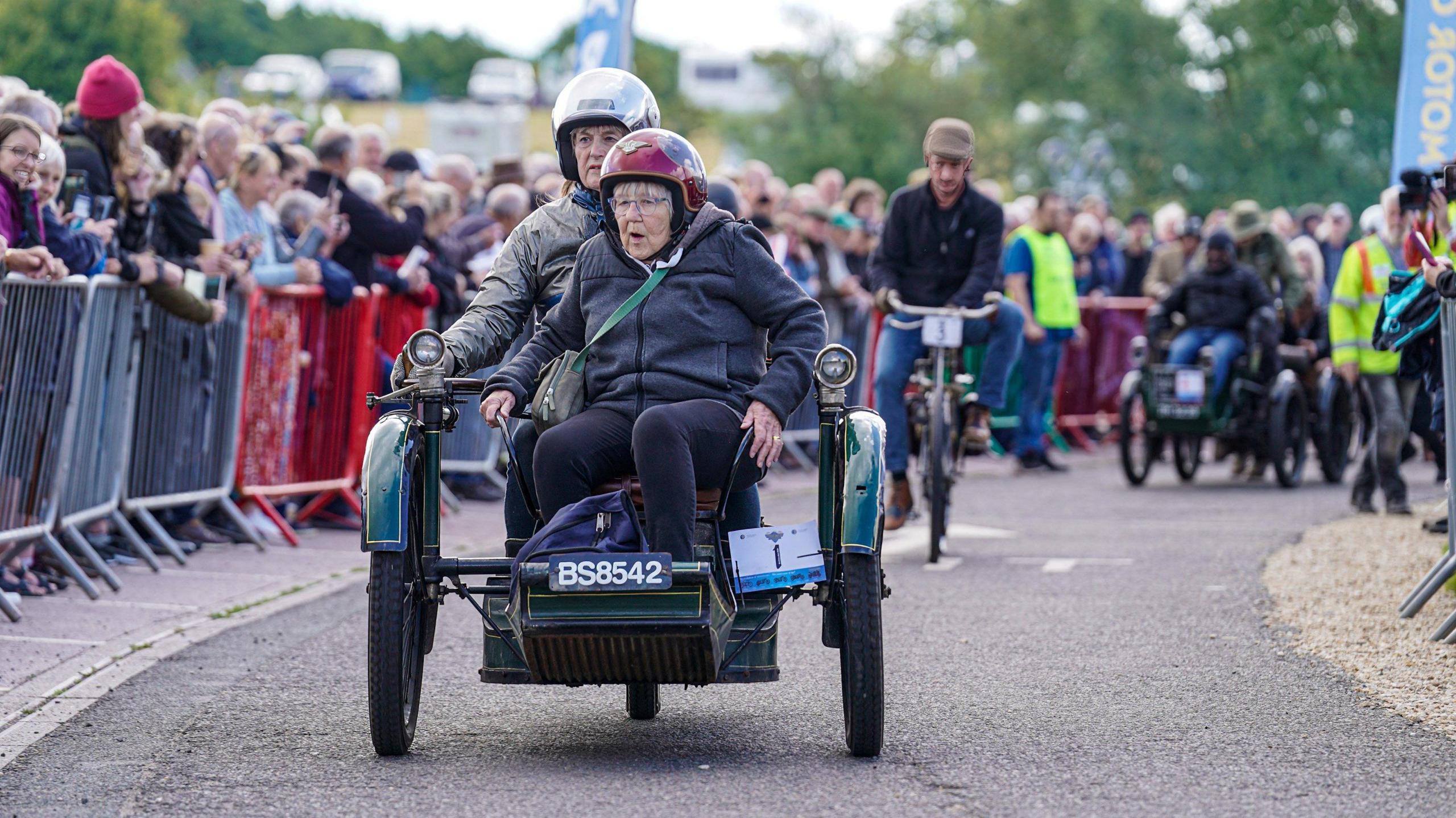 A vintage three-wheeled vehicle being ridden by a woman wearing a crash helmet. Sitting in front of her as a passenger is an elderly woman, also wearing a crash helmet and glasses. They are travelling along a road lined with crowds of people standing behind a barrier.