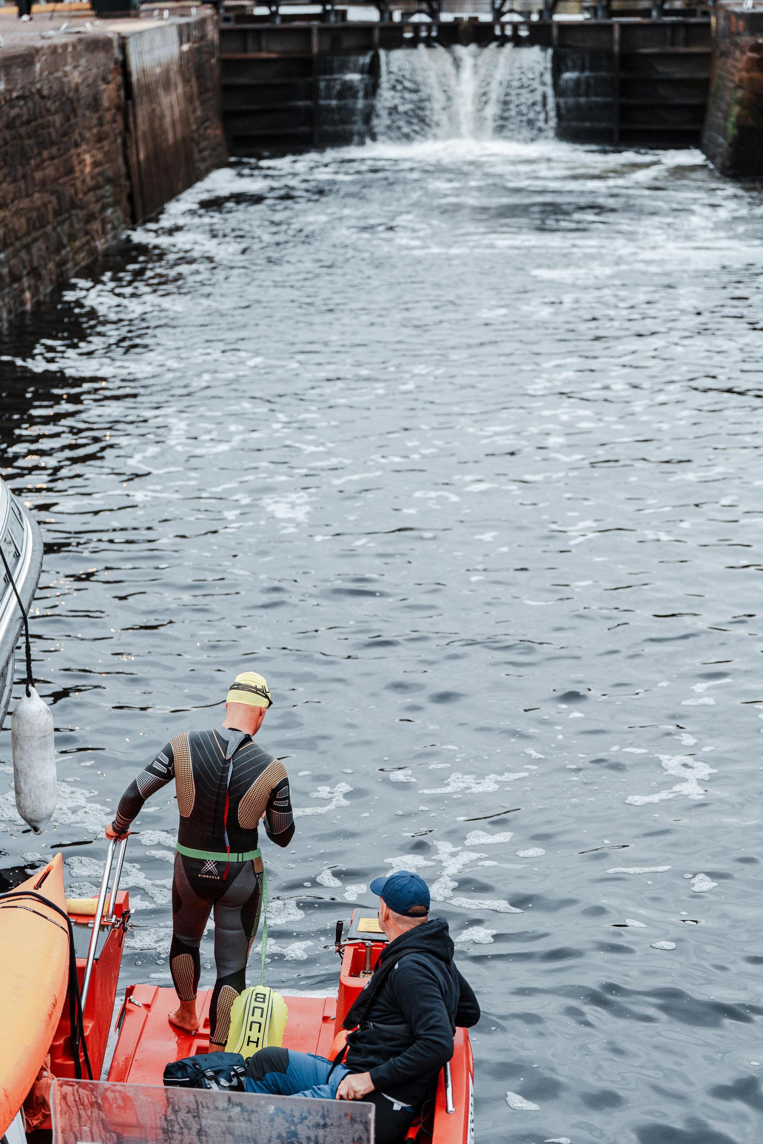 Iain is standing at a lock in his swimming gear along with a member of his support team. The water looks dark and cold.