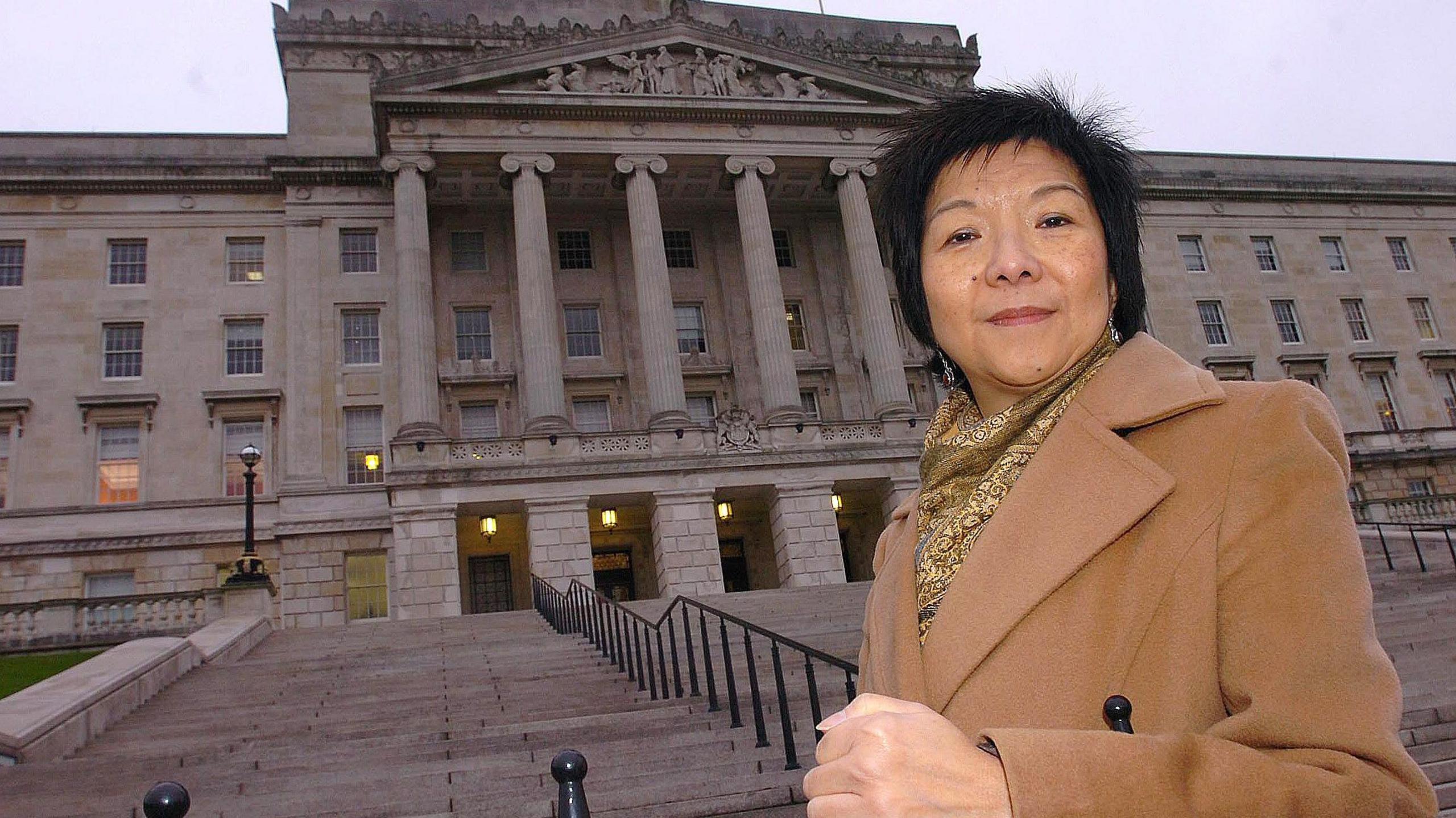 Anna Lo outside Parliament Buildings at Stormont.   She is looking to the camera and wearing a long beige coat and a gold scarf.