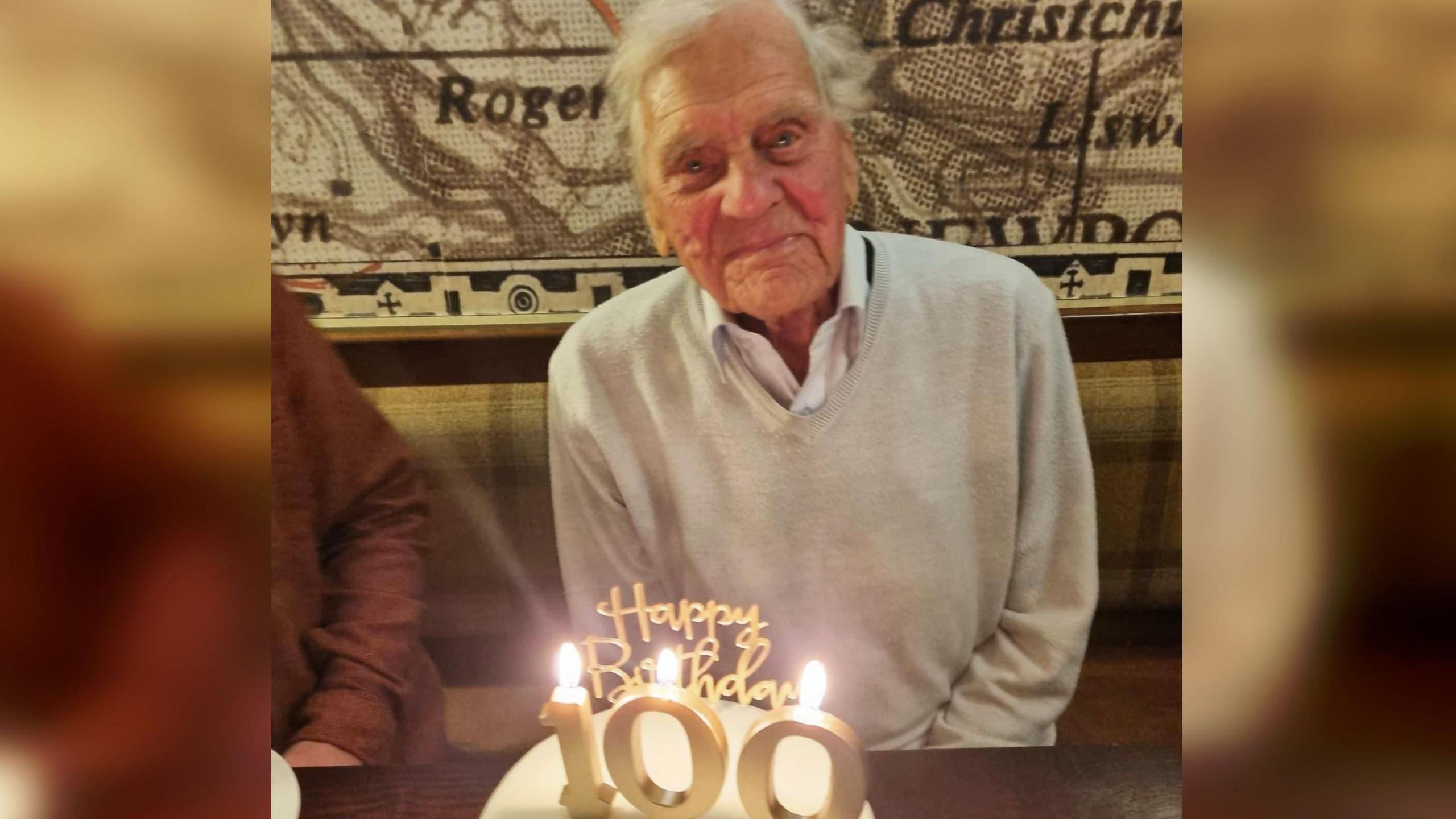 Phillip Ormerod yn dathlu ei ben-blwydd yn 100 oed - mae'n eistedd ger bwrdd ac o'i flaen mae cacen gyda chanwyllau a neges arni