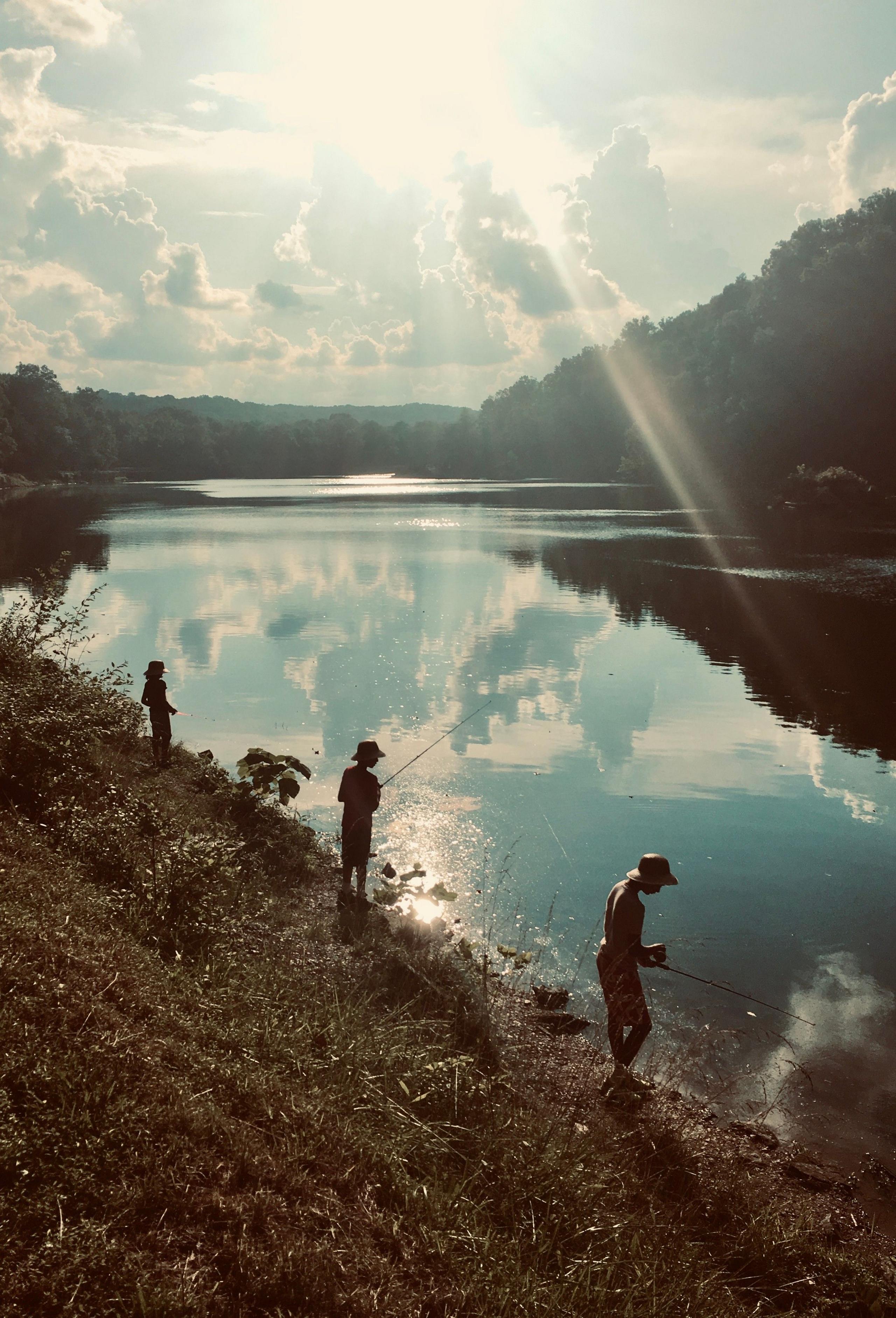 Three boys fishing on a river as the sun flares in the sky, with clouds reflected in the river