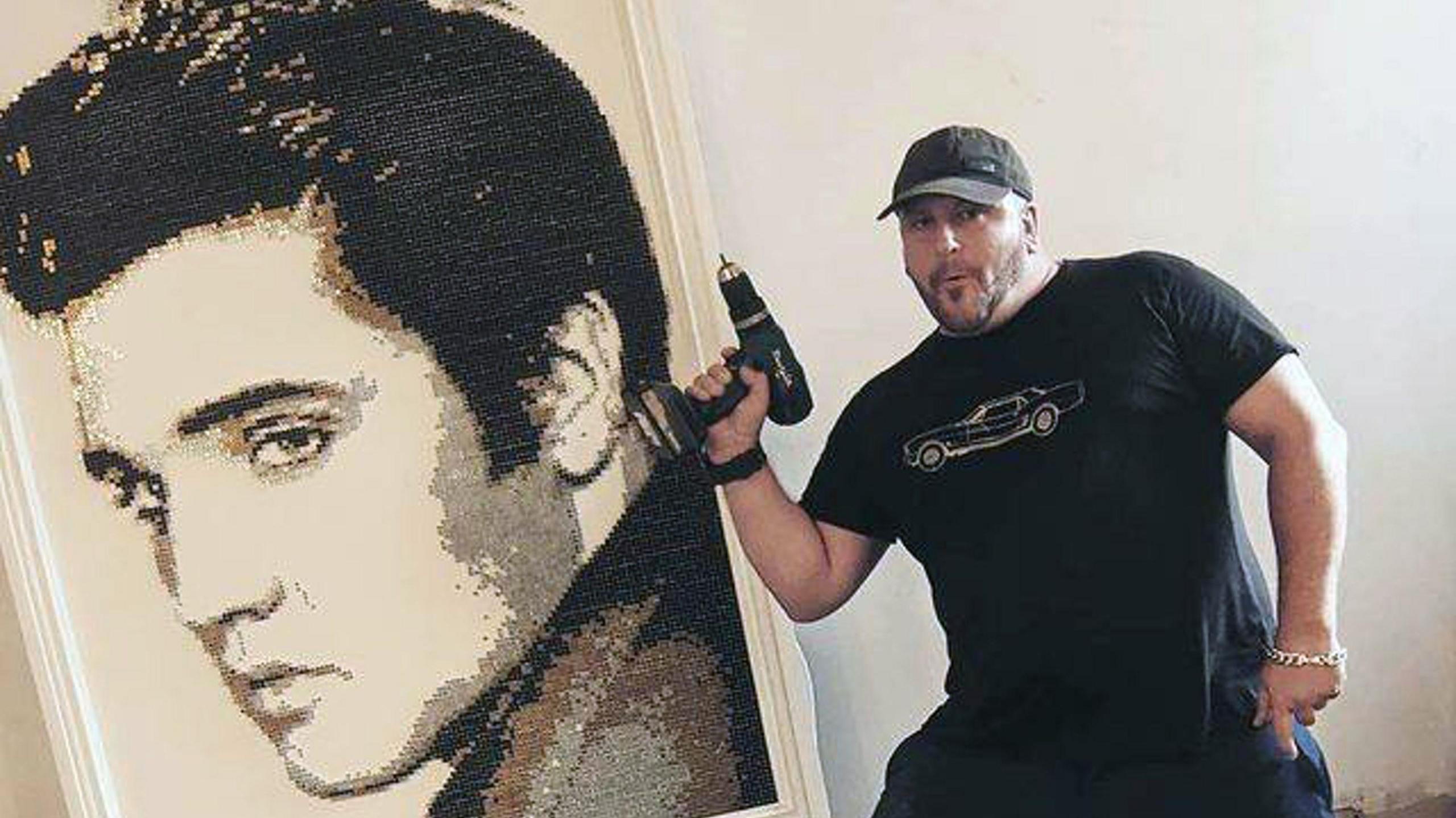 Darren Timby holding an electric screwdriver and standing next to a large portrait of Elvis made of screws.