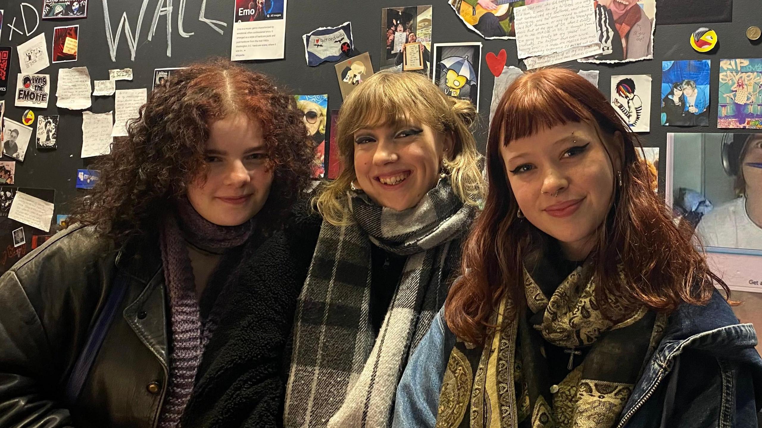 Three women - from left - Maddie, Seren and Molly standing in front of a display of small pictures and notes. Maddie has dark, curly shoulder-length hair and is wearing a scarf and leather jacket. Seren has dark blonde hair, with a checked scarf and coat and Molly has red hair, fringe and wears a denim jacket and paisley scarf.