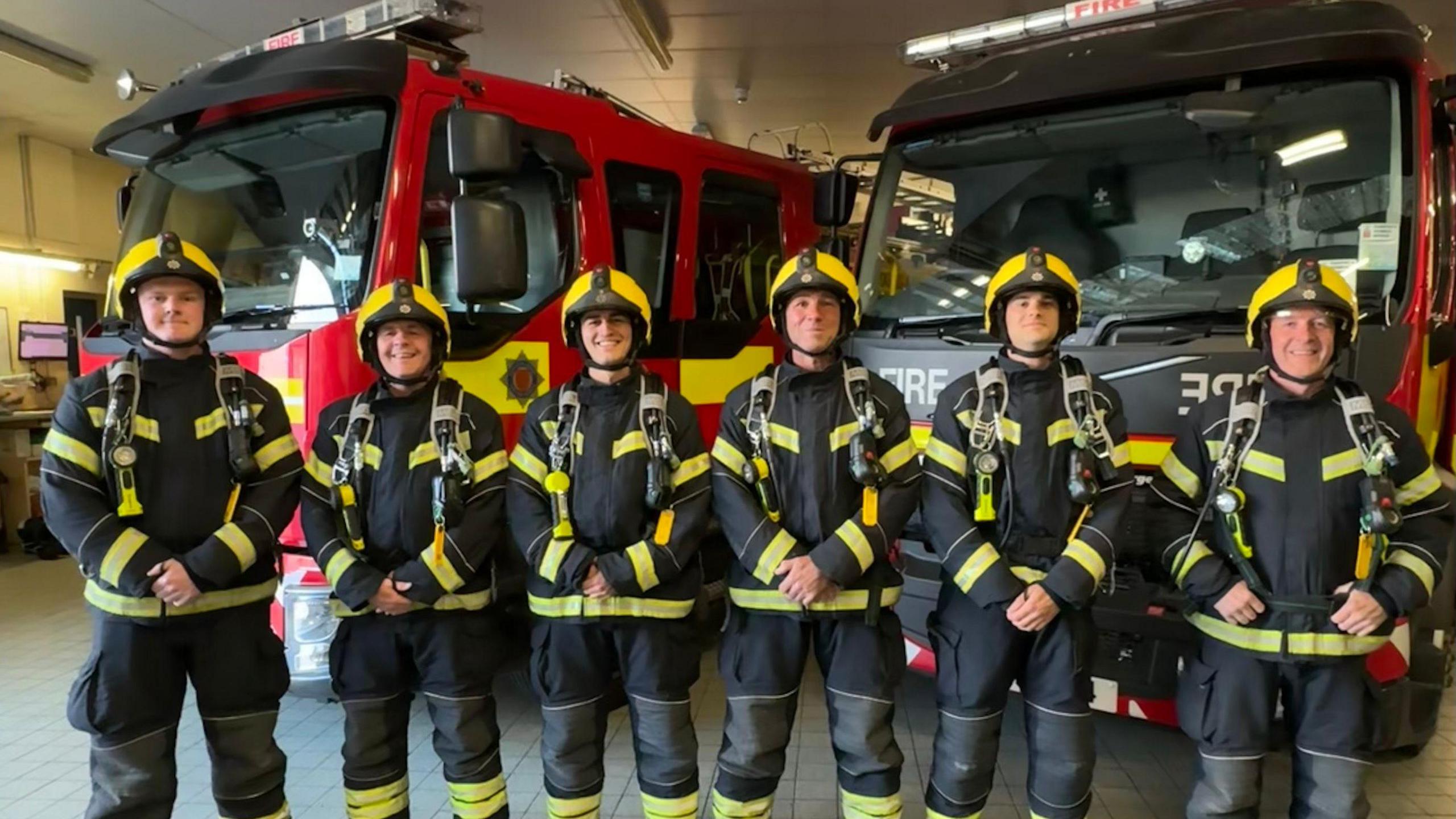 Six firefighters in full gear — helmets, jackets, pants, and boots — stand in a row inside a fire station. They face the camera with hands clasped in front. Behind them are two fire appliances, one red and one black with yellow stripes. The station’s interior, including ceiling lights, is visible in the background.