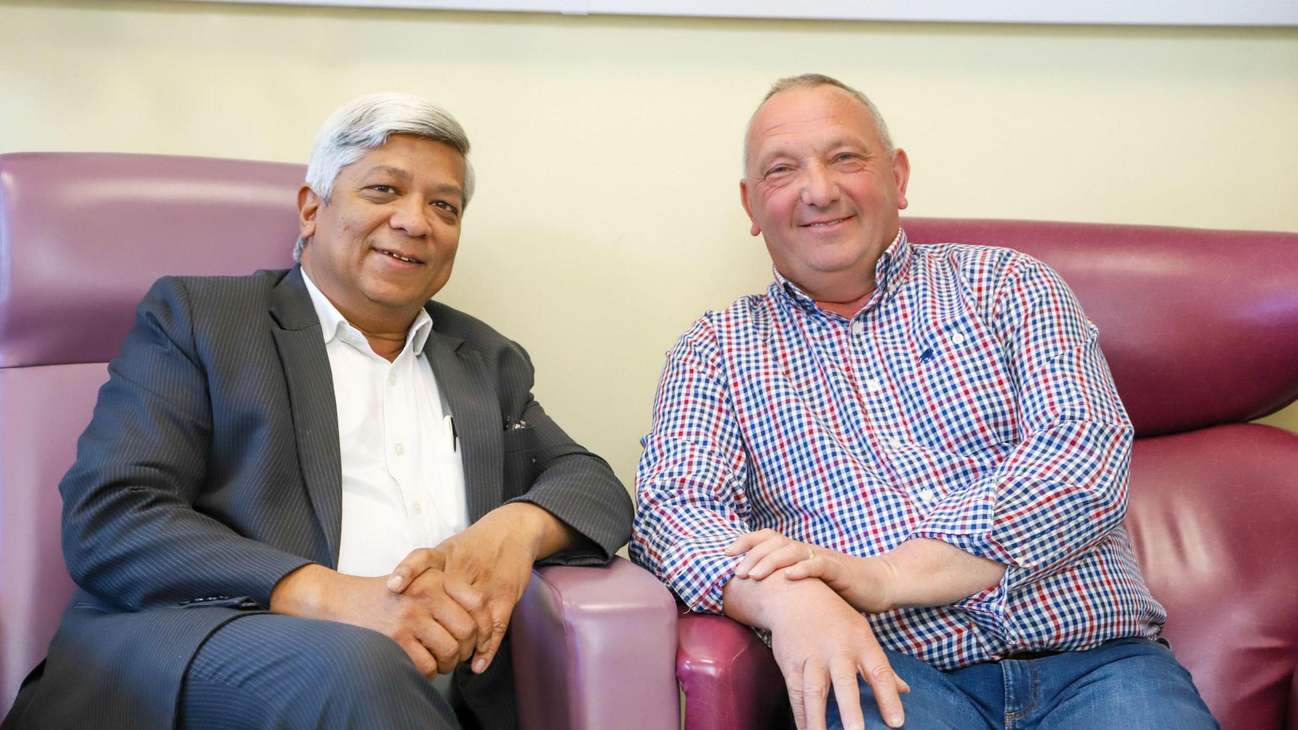 A man, wearing a checked shirt, and another man, wearing a black suit and white shirt. Sat in two purple chairs and smiling at the camera.