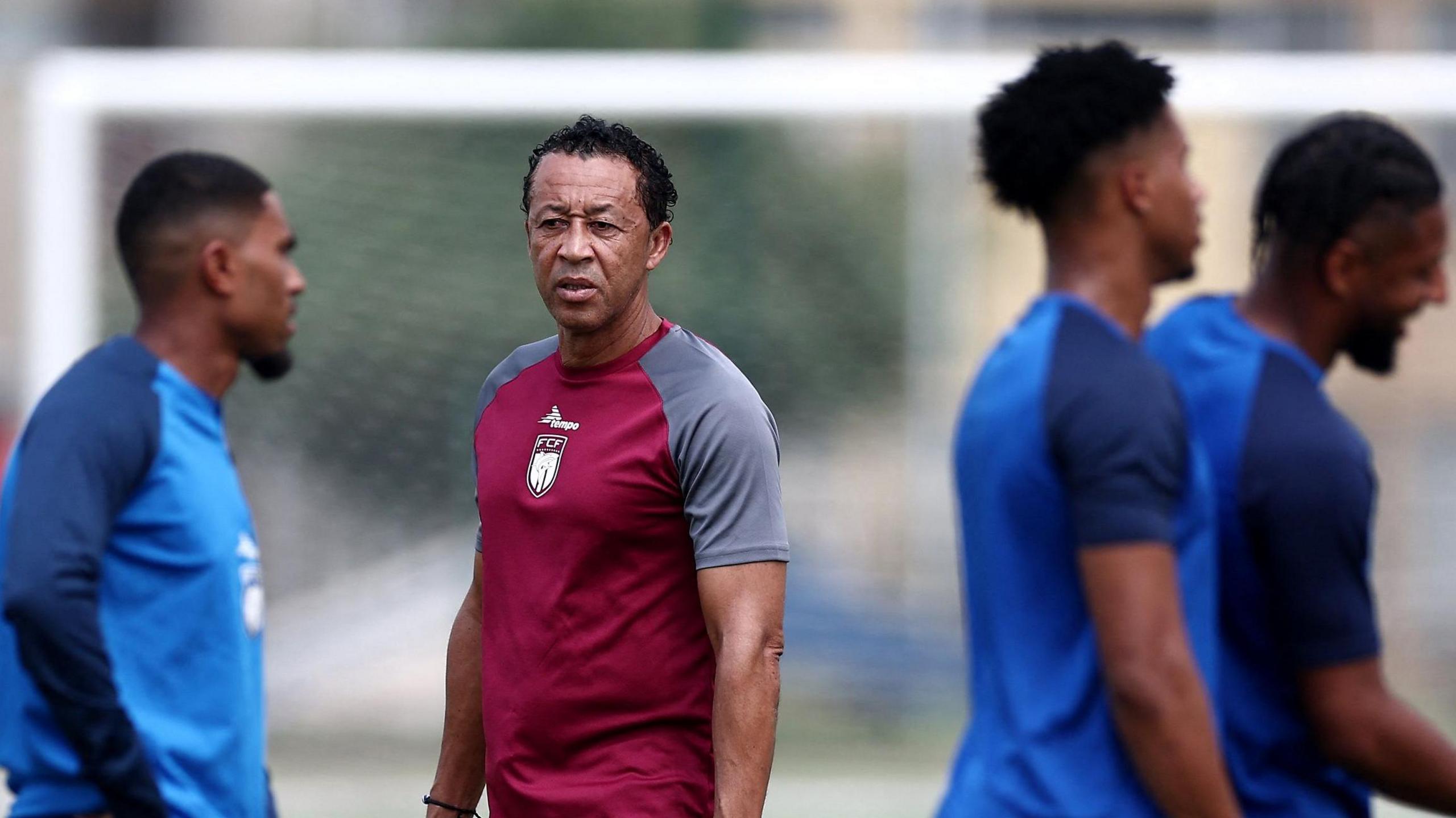 Cape Verde coach Bubista, wearing a maroon training top, stands during a training session overseeing three players in blue shirts who are out of focus in the foreground