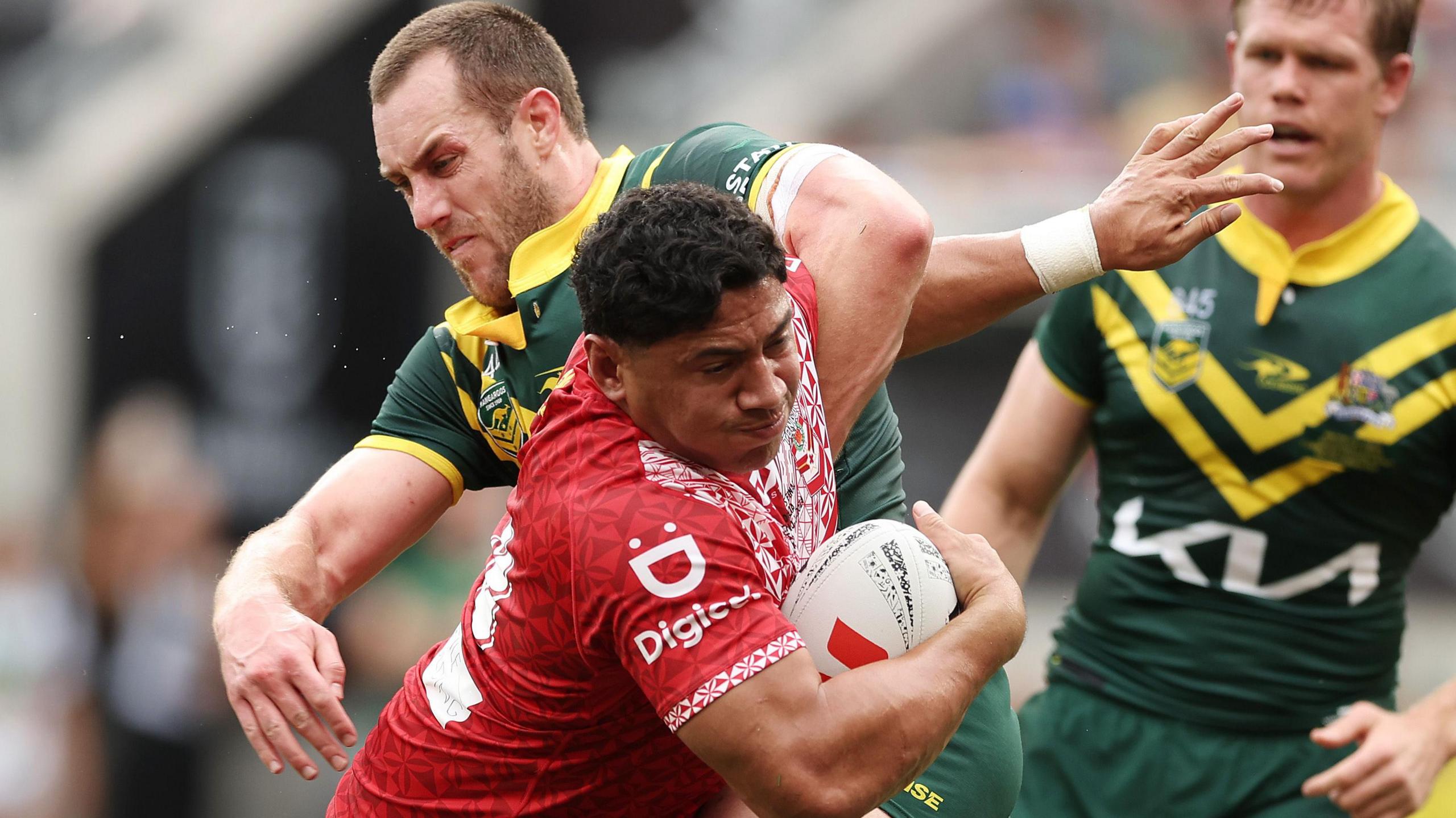 Isaah Yeo for Australia in green and gold kit puts a wrestle-style tackle on Tonga's powerful Jason Taumalolo in a Pacific Championship match in 2024 while Yeo's teammate Lindsay Collins looks on from the background.