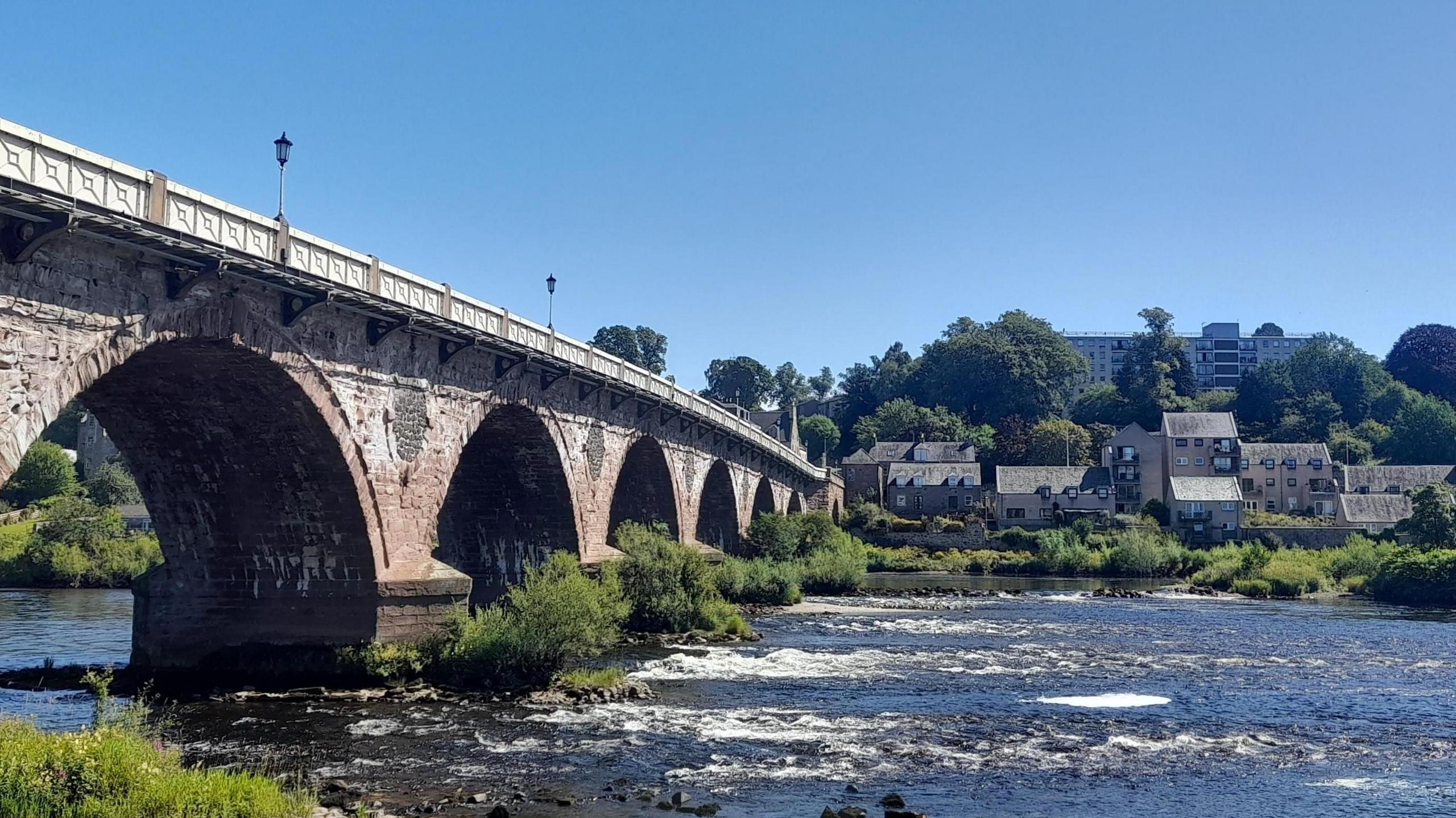 A bridge over a river with some buildings on the far bank under a blue sky