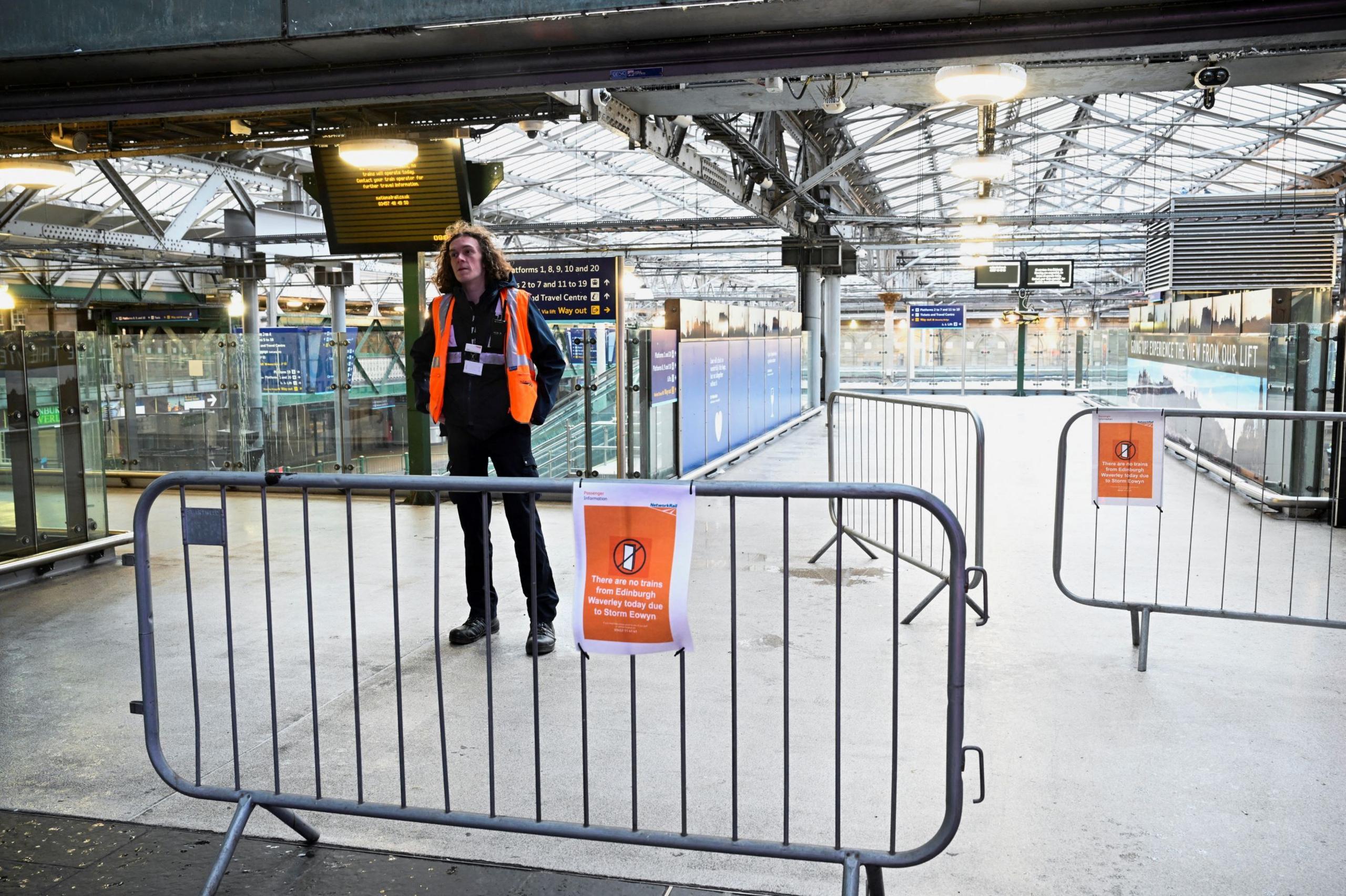 A guard with long curly brown hair and an orange tabard stands alone in the station concourse. There are signs and barriers everywhere saying the trains are not running.