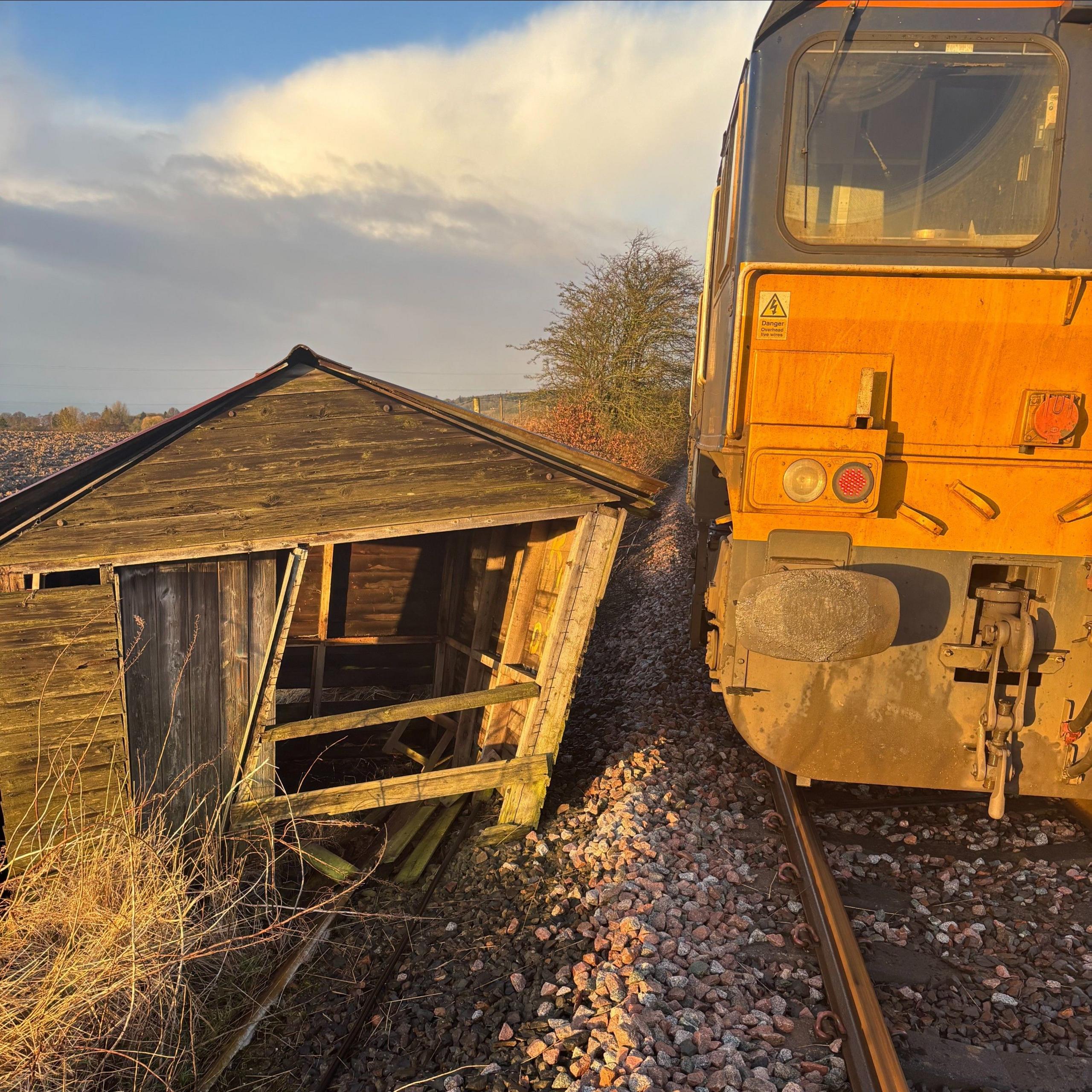 A very battered looking old garden shed stands just on the edge of a railway  track in the countryside. A diesel engine stands right beside it. It is sunny and the sky is blue.