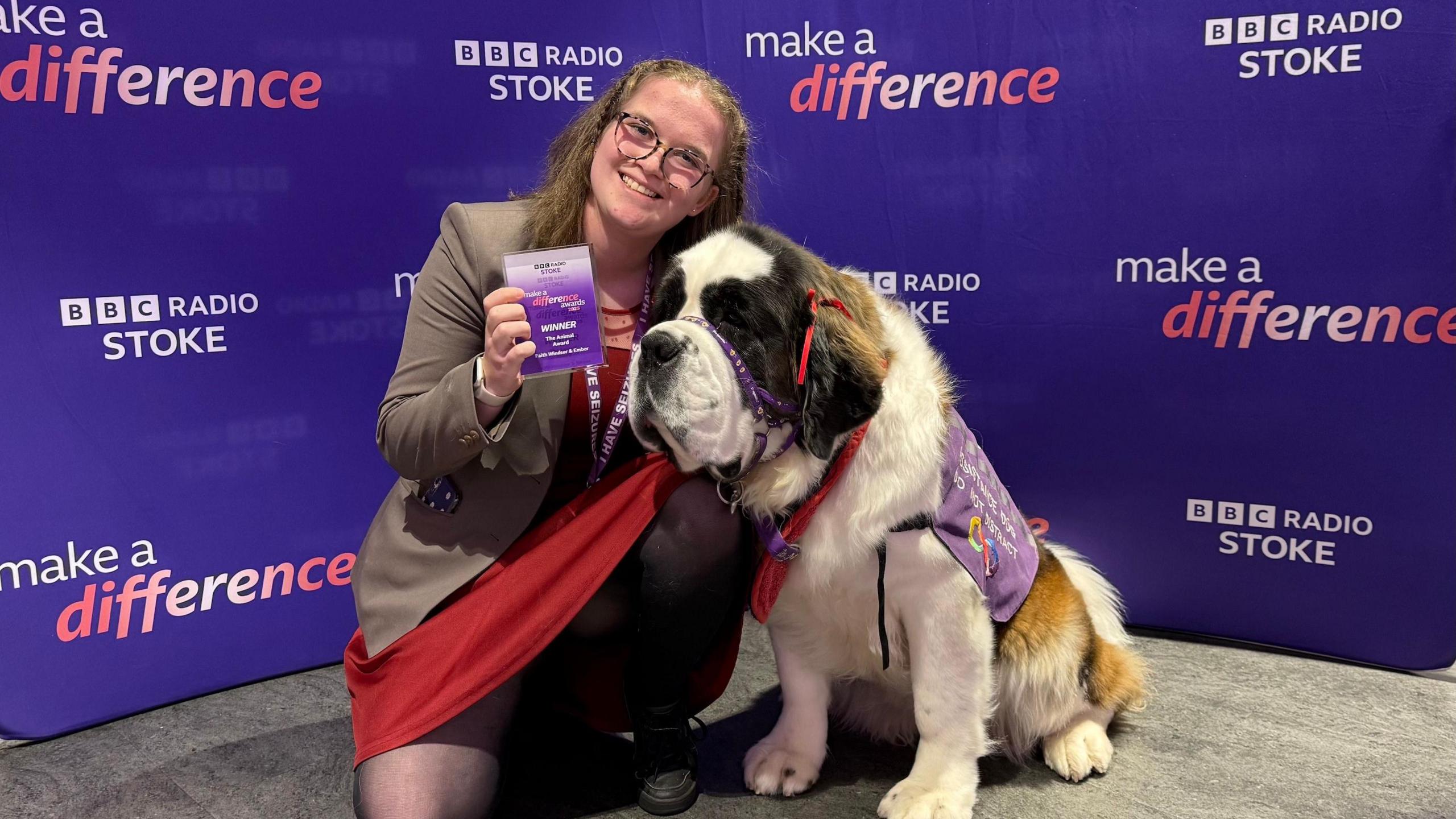 Faith Windsor is crouching in front of a purple BBC Radio Stoke Make A Difference step and repeat board next to her dog Ember. Faith is wearing a red dress and cream-coloured jacket. Ember is wearing a purple dog jacket which identifies her as an assistance dog.