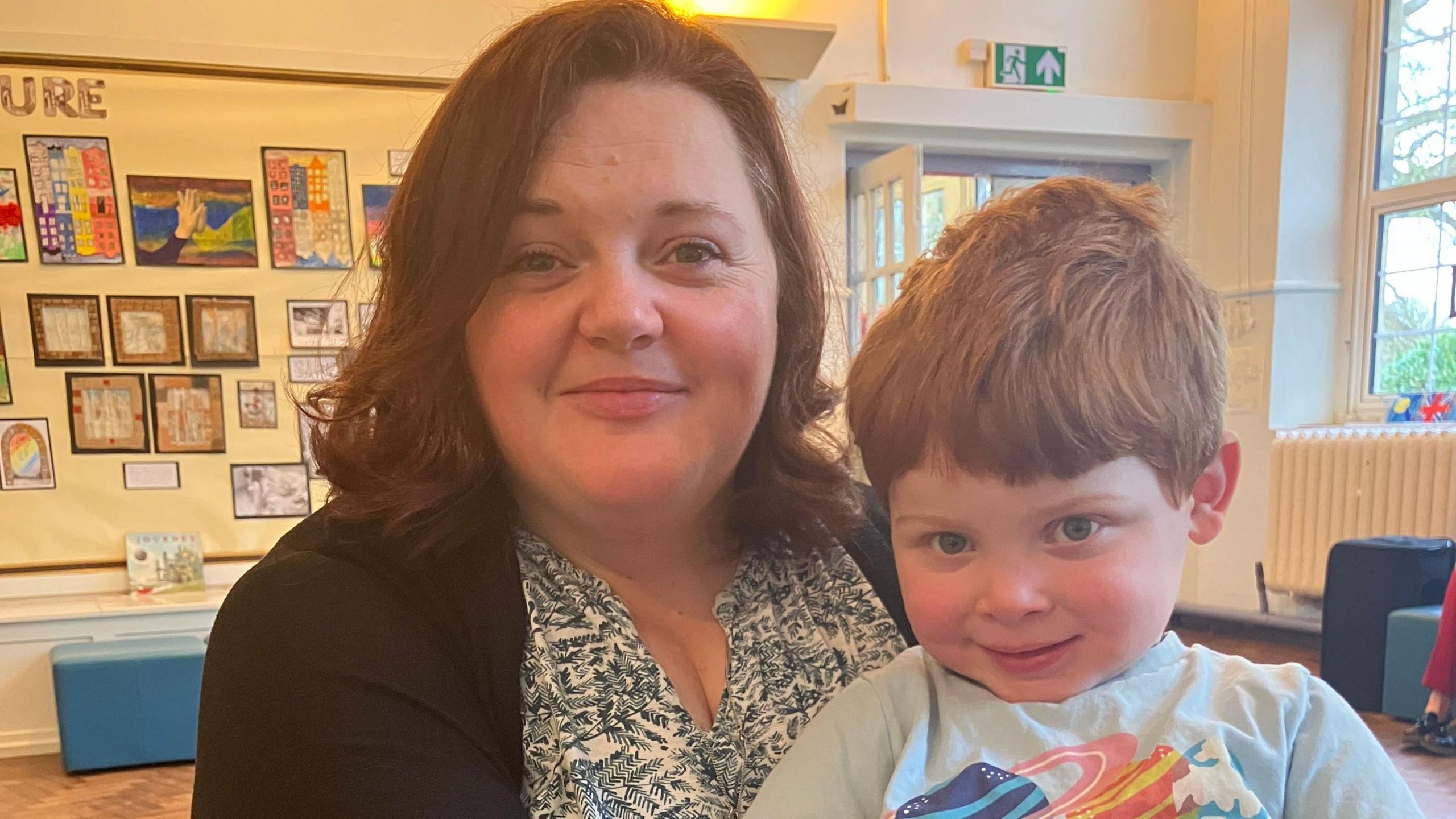 Kathryn Davidson is sitting in a classroom with her son Rory on her knee. She is wearing a shirt with leaves on and a black cardigan and Rory is wearing a blue t-shirt. They are both smiling.