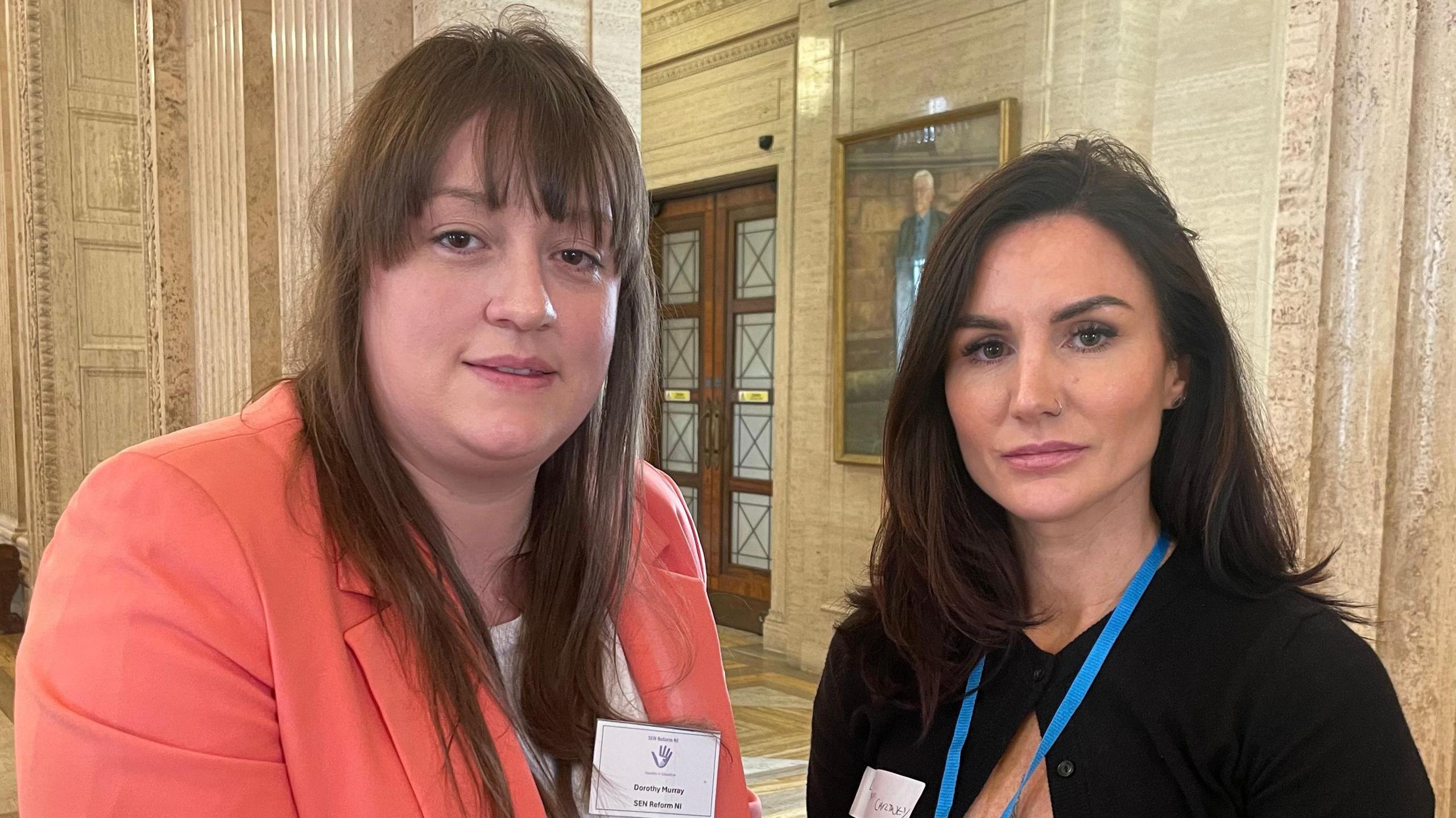 Two woman standing next to each other. the woman on the left is wearing a salmon blazer and white top, the woman on the right is wearing black. Both women have  brown hair and are wearing blue lanyards