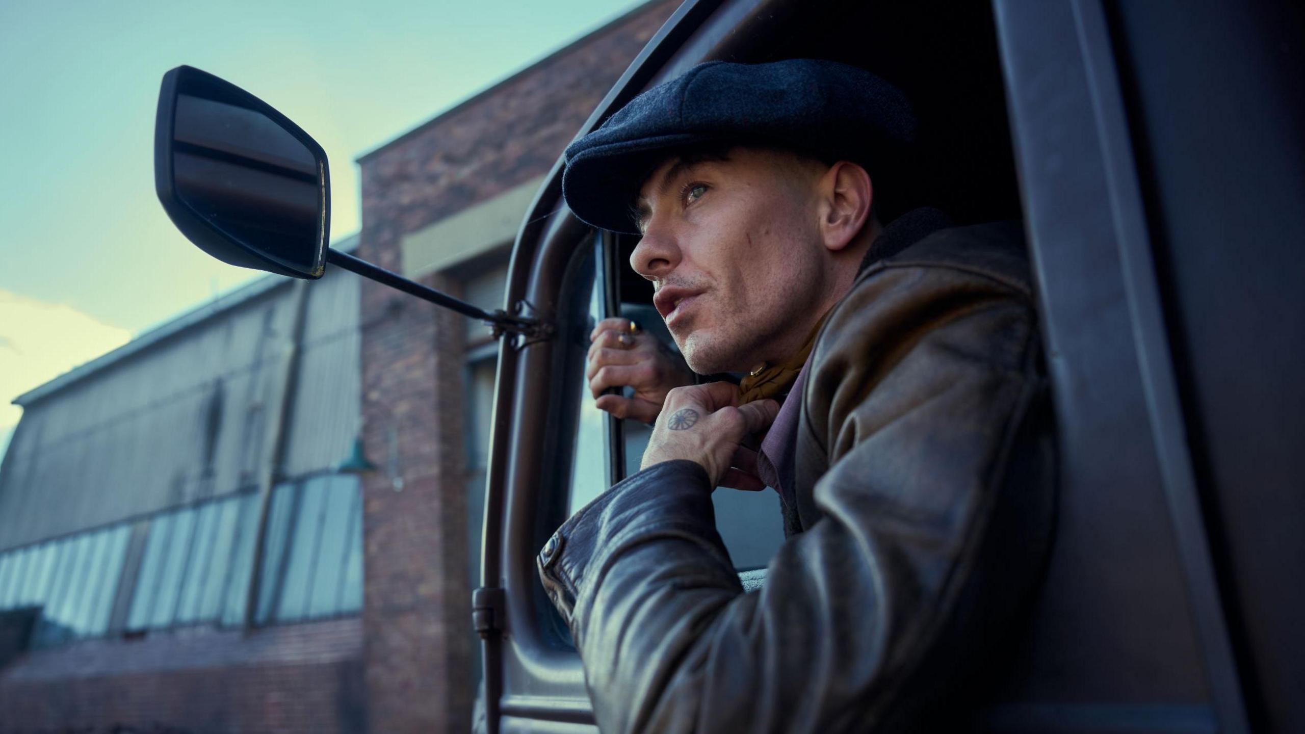 Barry Keoghan wearing a flat cap and leaning out the side of a car as he pulls up to an industrial building.