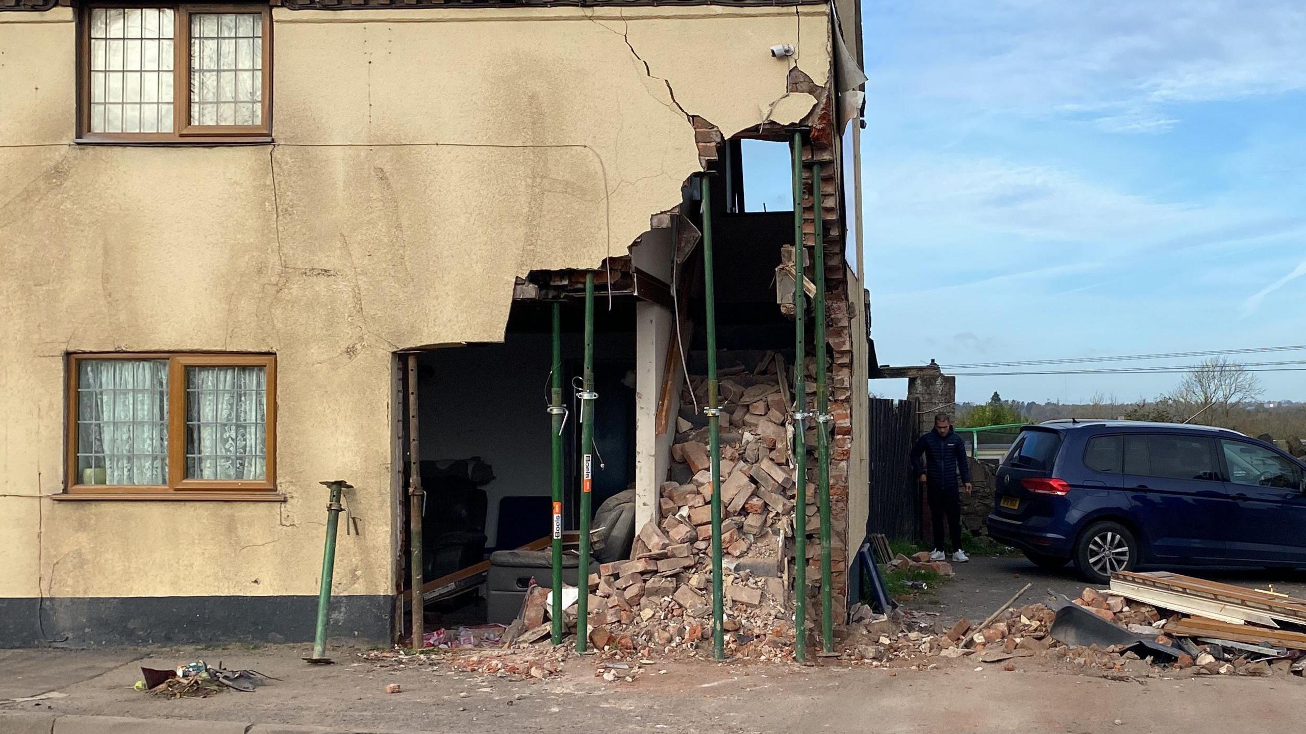 A damaged building with beige walls, with a large chunk missing from the side and lots of exposed bricks. The walls are propped up with green metal poles and there is a blue car parked alongside