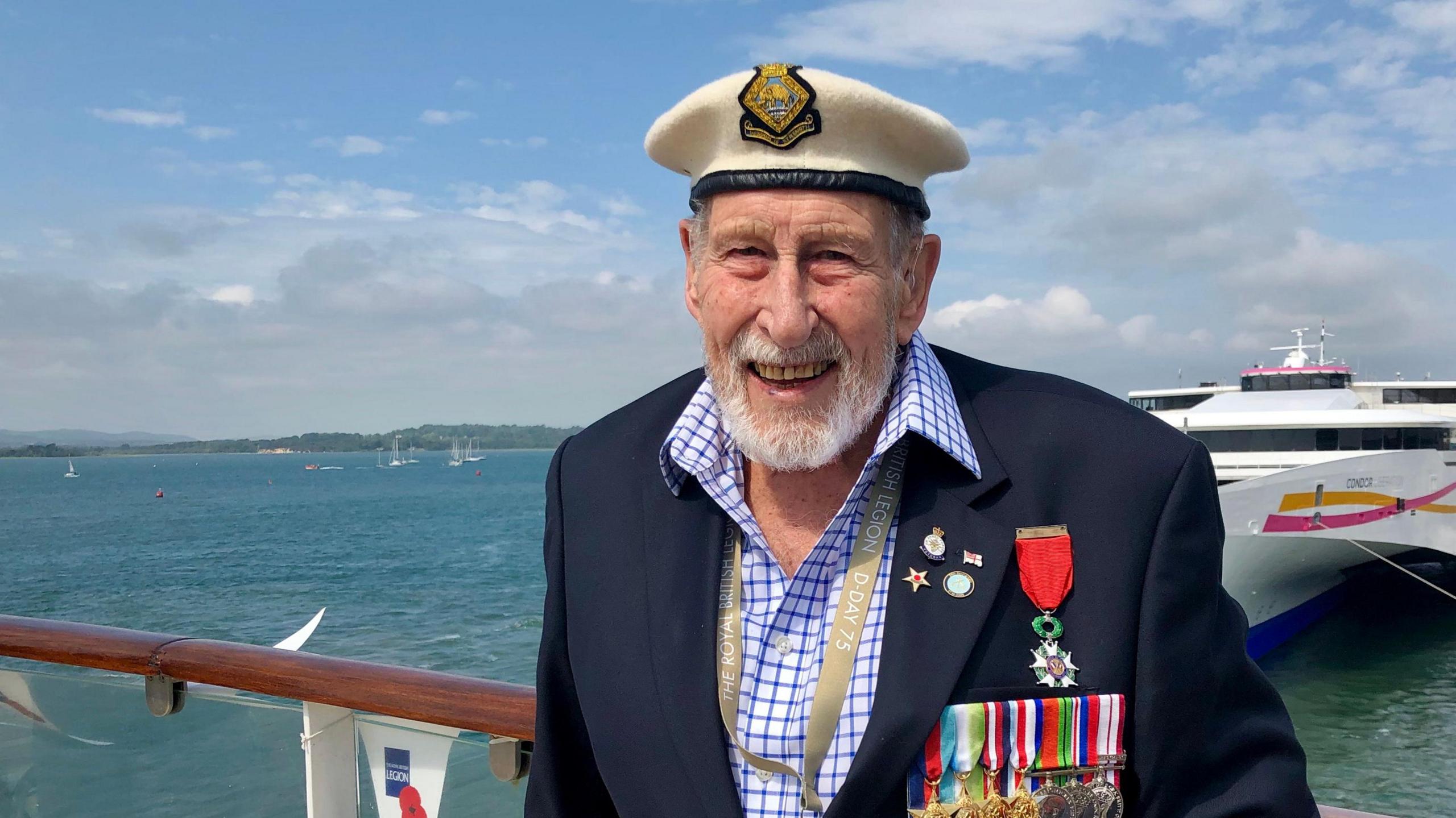 George Winter from shoulders up wearing a suit adorned in medals and wearing a sailors hat. He is stood on a boat, with other boats in the background as well as the water and shoreline in the distance.