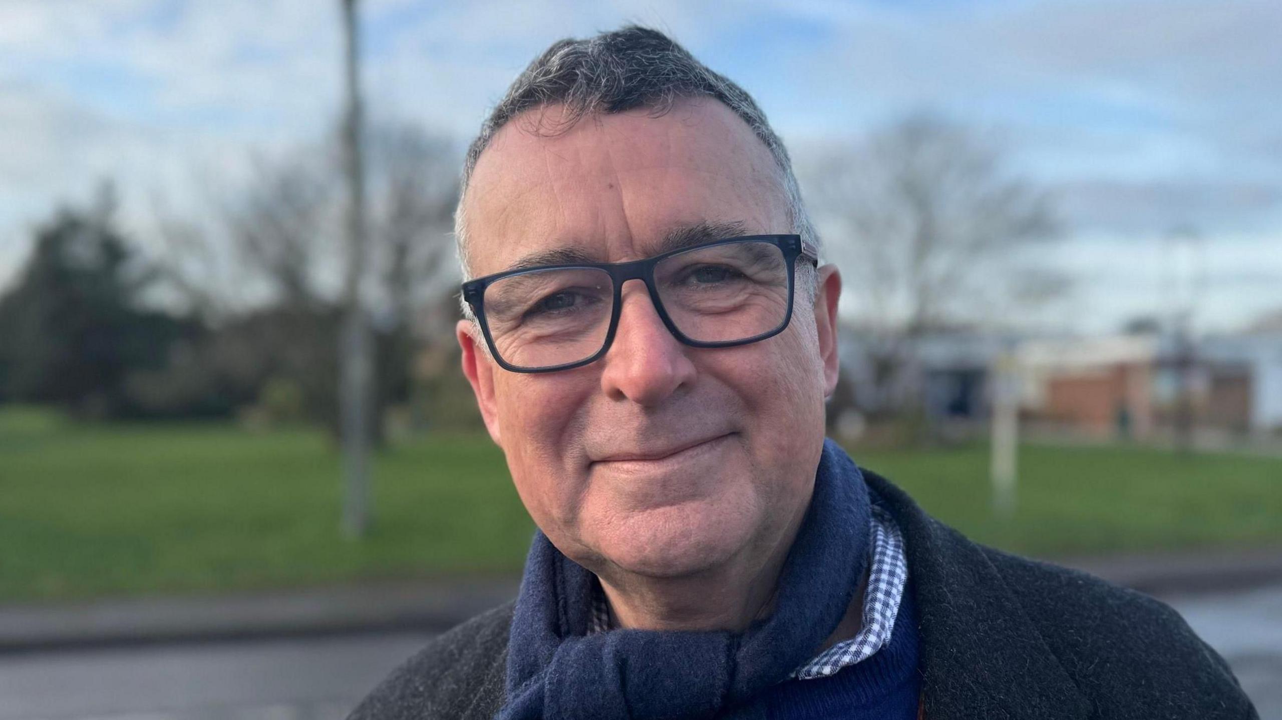 Sir Bernard Jenkin smiling straight at the camera. He is standing on a pavement opposite a community centre and is wearing a navy scarf, dark grey coat, and a royal blue jumper over a blue and white checked shirt.