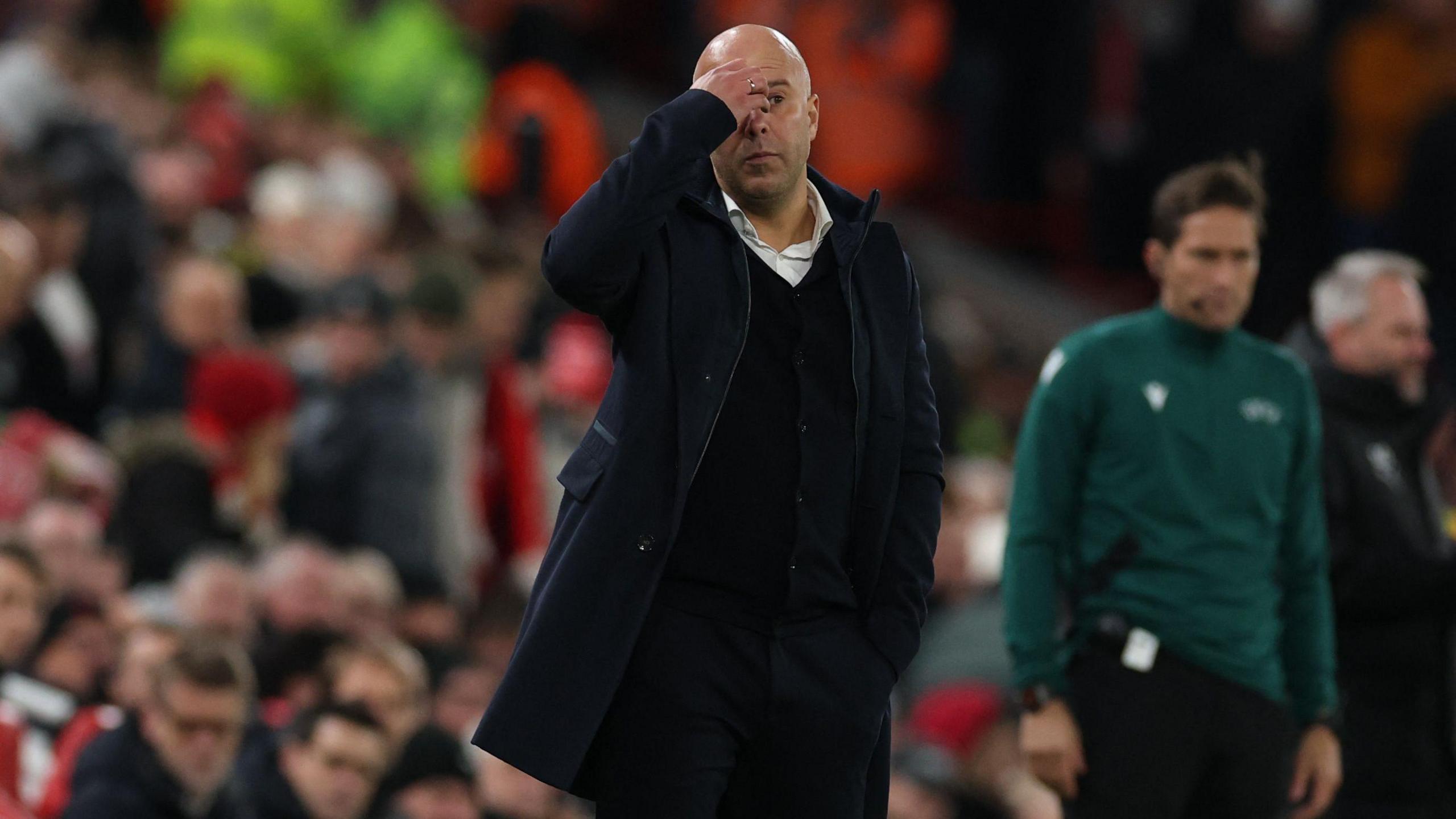 Liverpool manager Arne Slot stands in front of a crowd of fans. He wears a dark coat and cardigan with a white shirt, his hand rests on his forehead in frustration. 