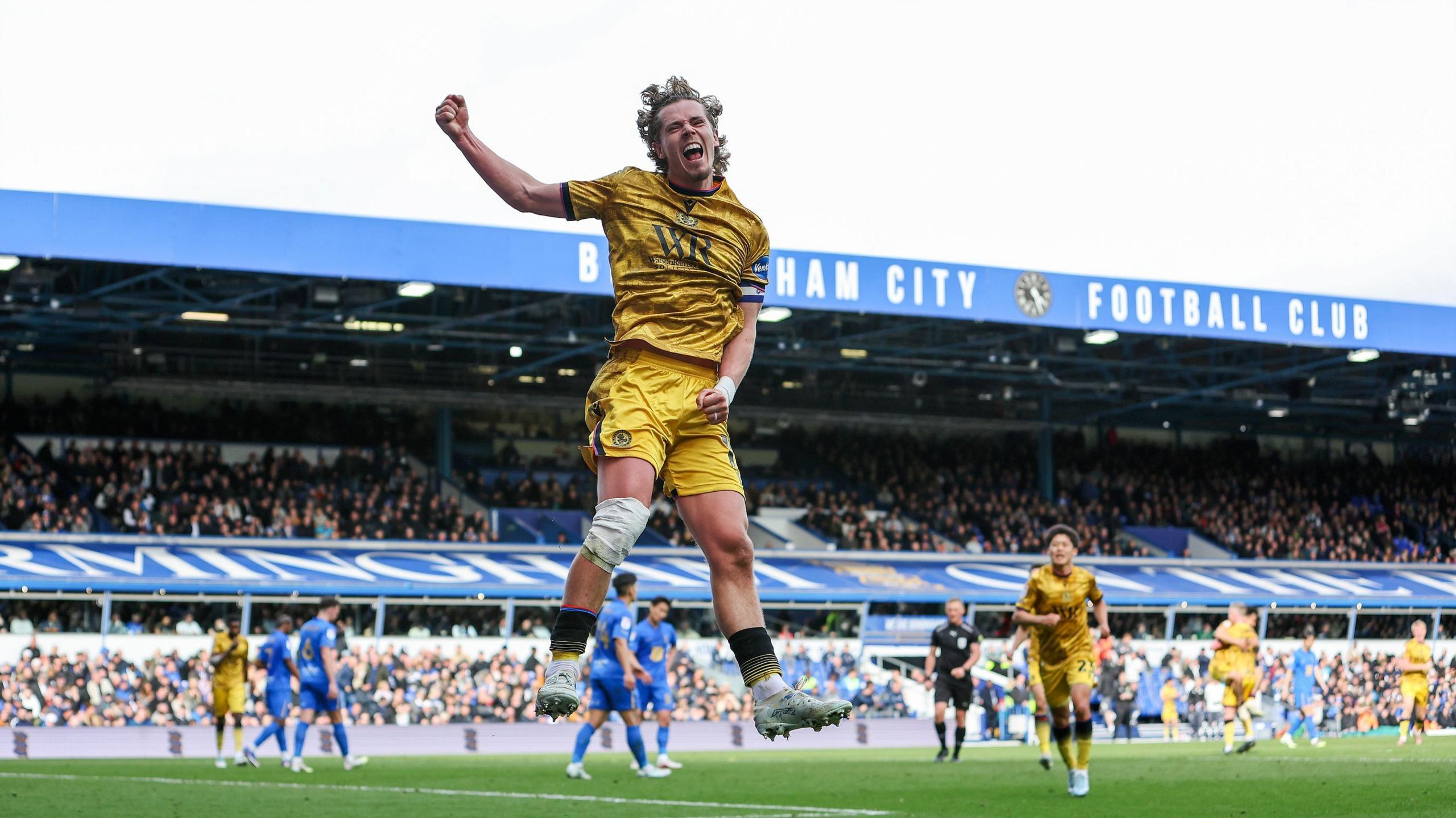 Todd Cantwell leaps into air celebrating, right harnd above his head, yelling at fans, wearing away kit