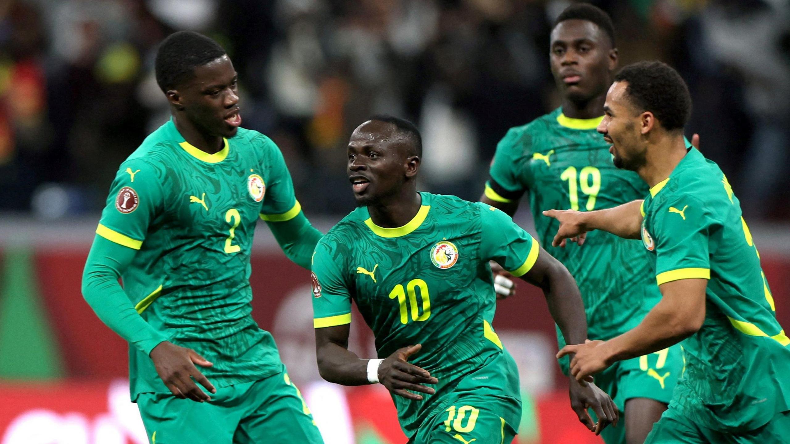 Sadio Mane, wearing a green Senegal strip with yellow detail and a number 10 on the chest, wheels away in celebration as he is chased by three team-mates
