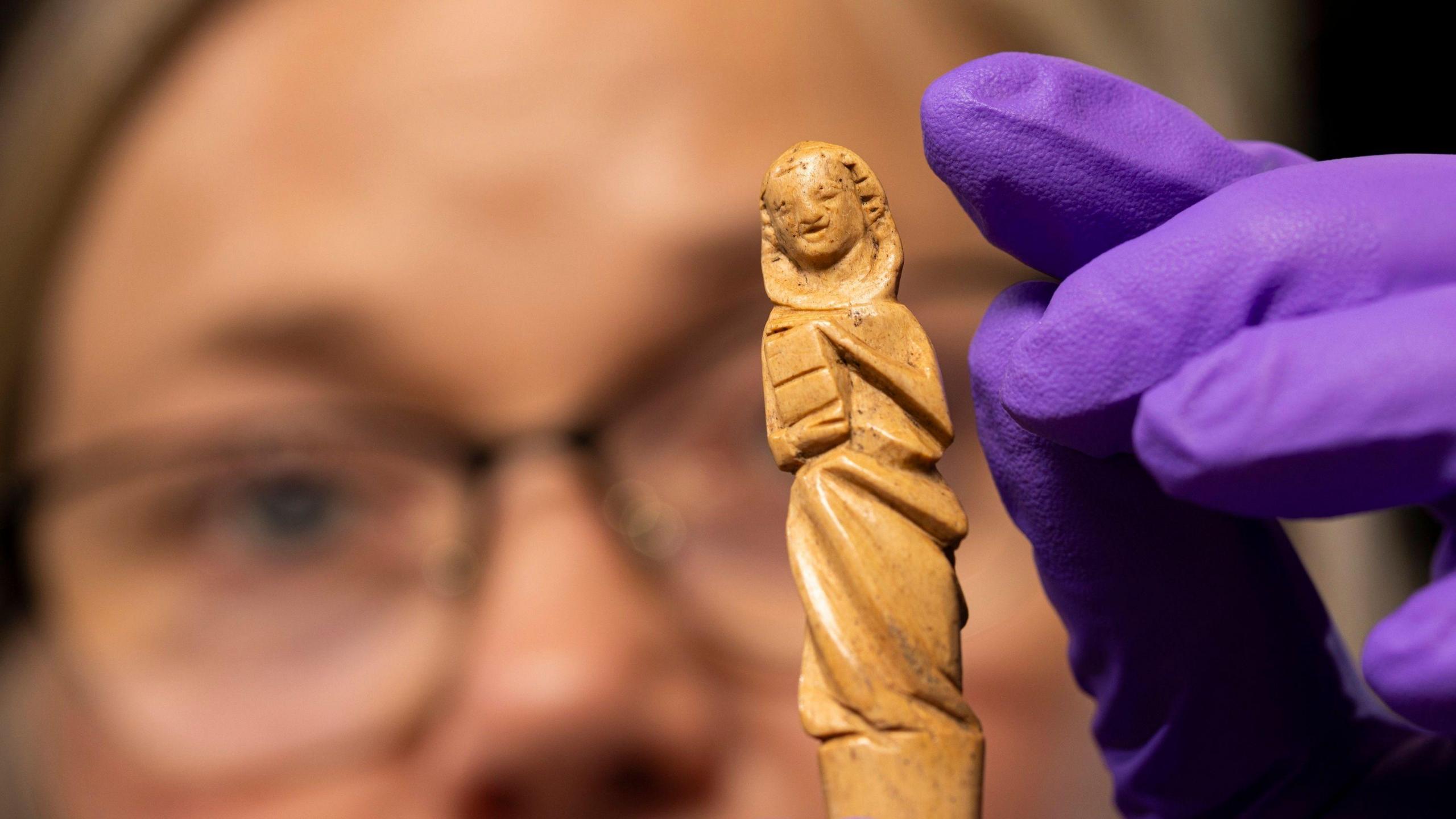 A close up of the gravoir, an ornately carved piece of red deer antler. The carving is of what looks like a hooded figure holding a book. Dr Alice Blackwell is holding the gravoir in protective purple gloves. Dr Blackwell's face is just out of focus.
