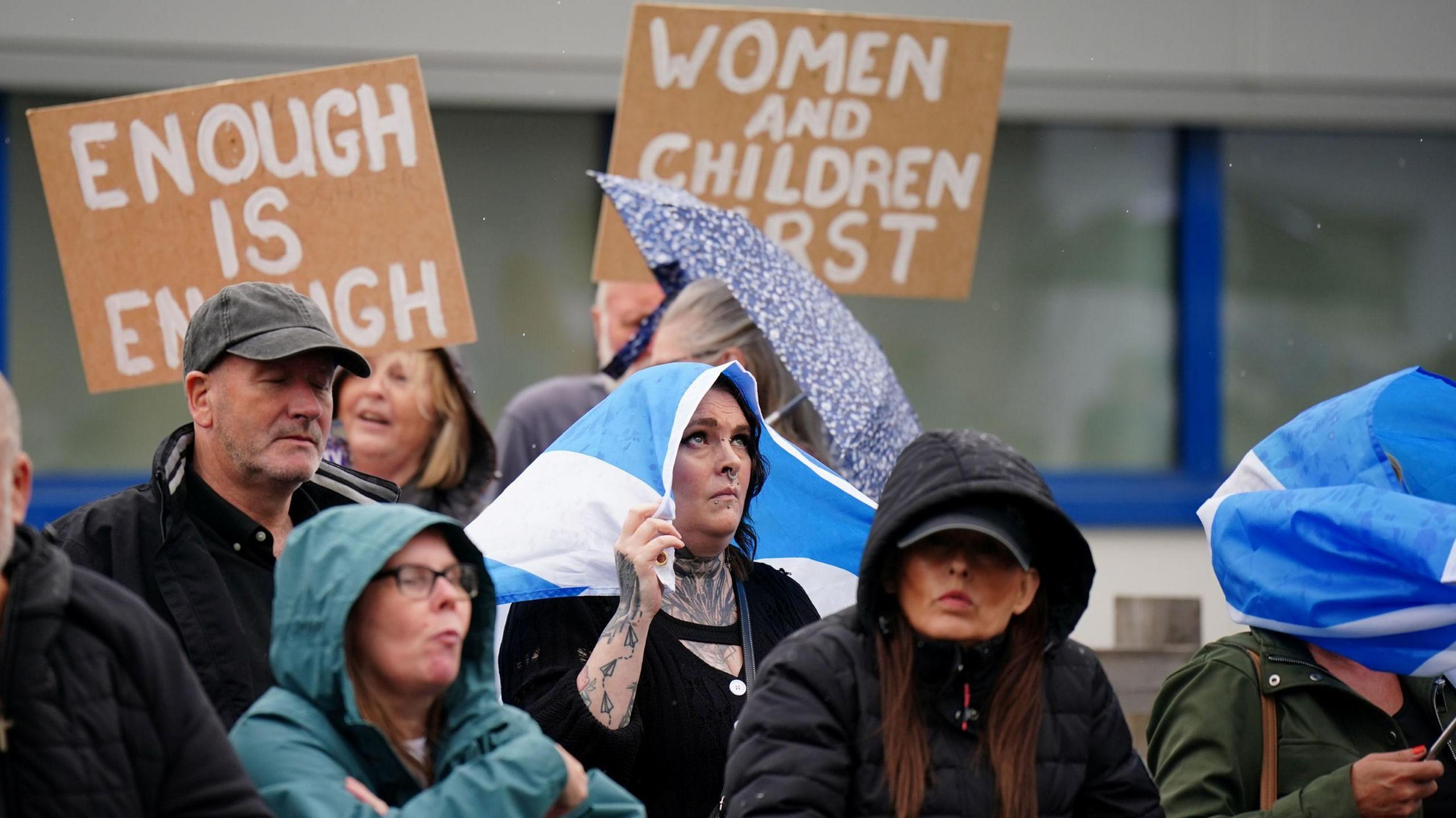 A group of women stand together. Women in the crowd have their hoods up, while one shelters from rain under a Saltire flag. Two signs with words painted on brown boards are in the background. One reads: "Enough is Enough" and the other: "Women and children first".