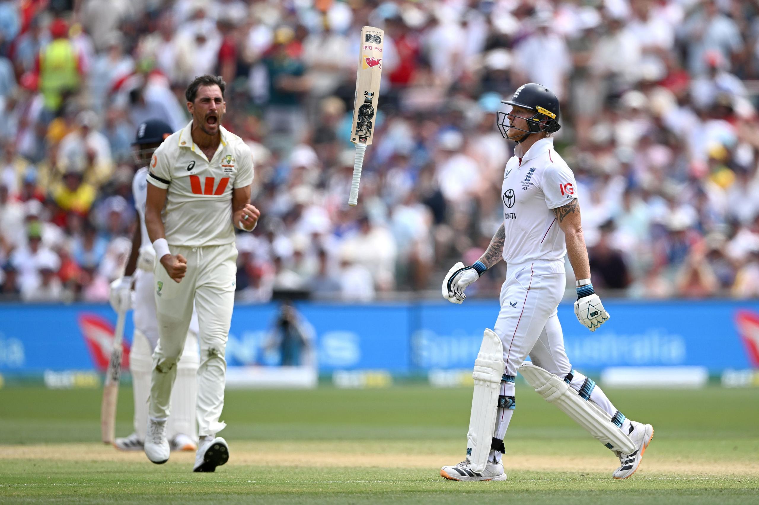 A cricket bat is seen mid-air between two players during a tense moment on the field. One player in white bowling attire reacts energetically, while the batter in white gear and a dark helmet walks forward, holding their gloves up. A packed stadium crowd forms a blurred backdrop to the action.