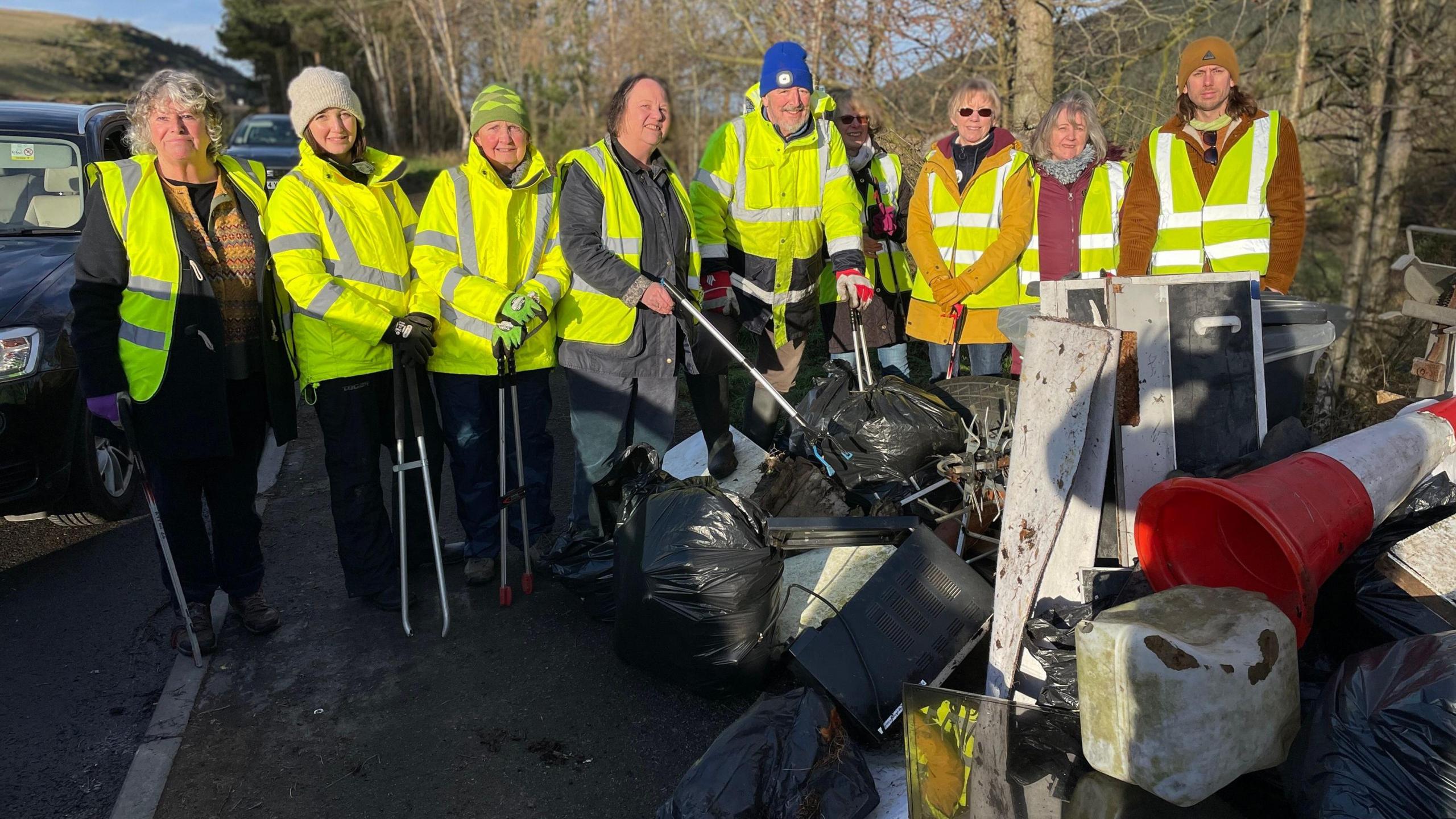 A group of nine people in hi-vis jackets with around a dozen black rubbish bags in front and other items of waste