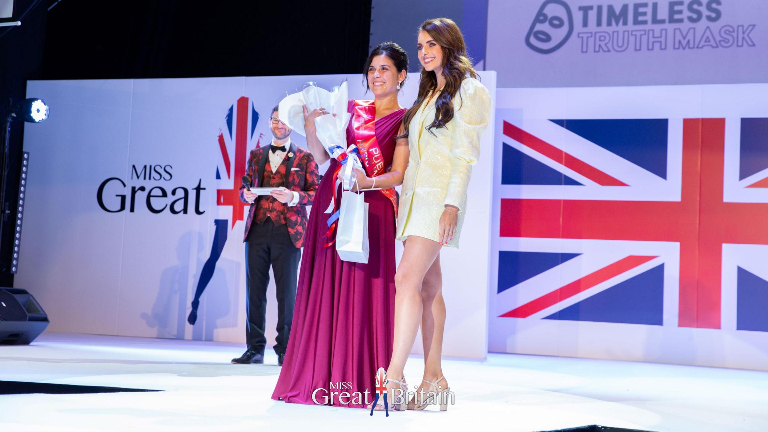 Elle Seline in a long pink dress, smiling, holding a bouquet of flowers. There is a Miss Great Britain backdrop on the stage she is standing on. There is a woman in a white shimmering jacket/dress standing next to her and a man in a red and black suit in the background, holding cue cards.