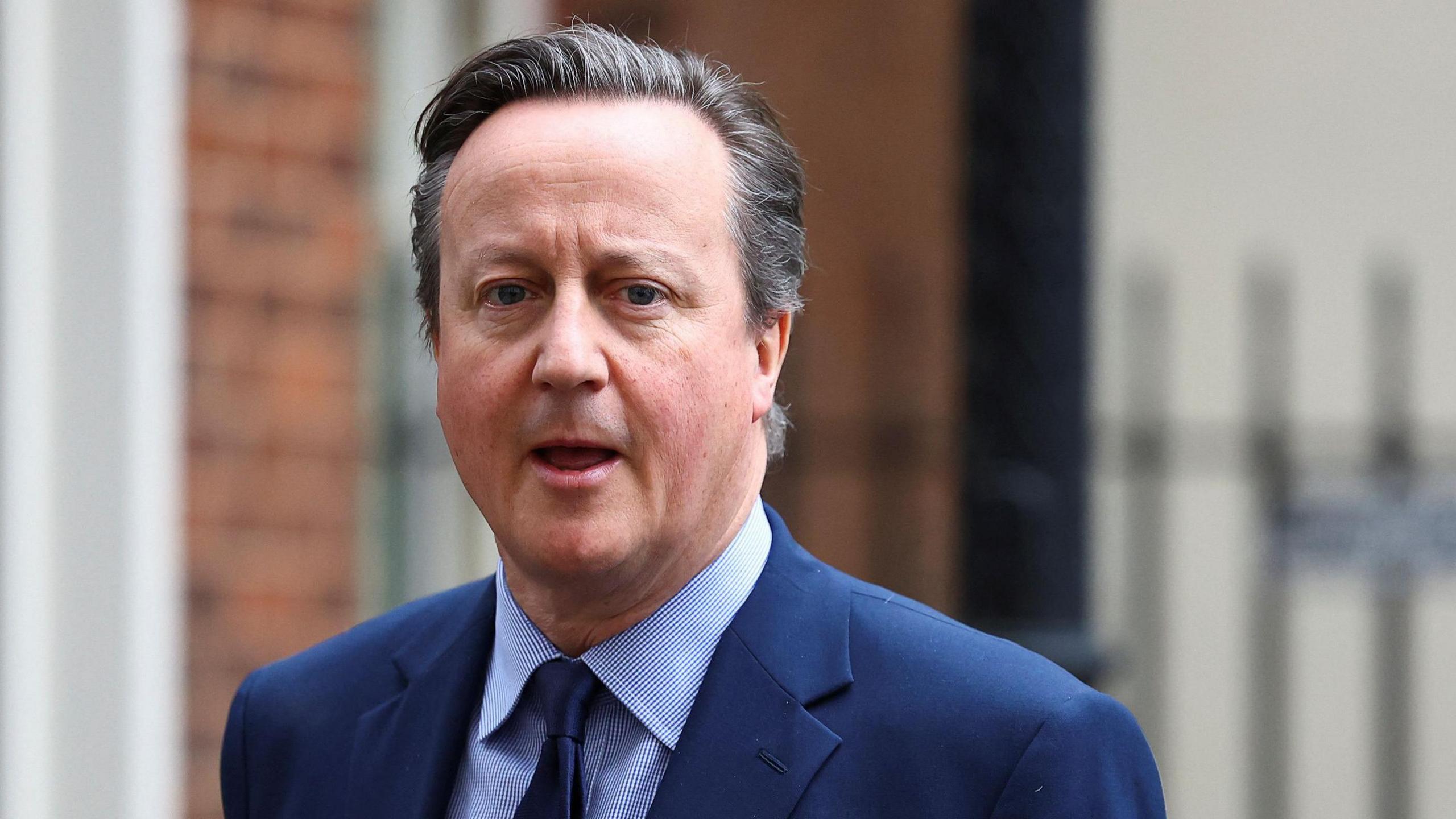David Cameron walking out of Downing Street in London. He is wearing a blue blazer, blue tie and a shirt with blue and white stripes.