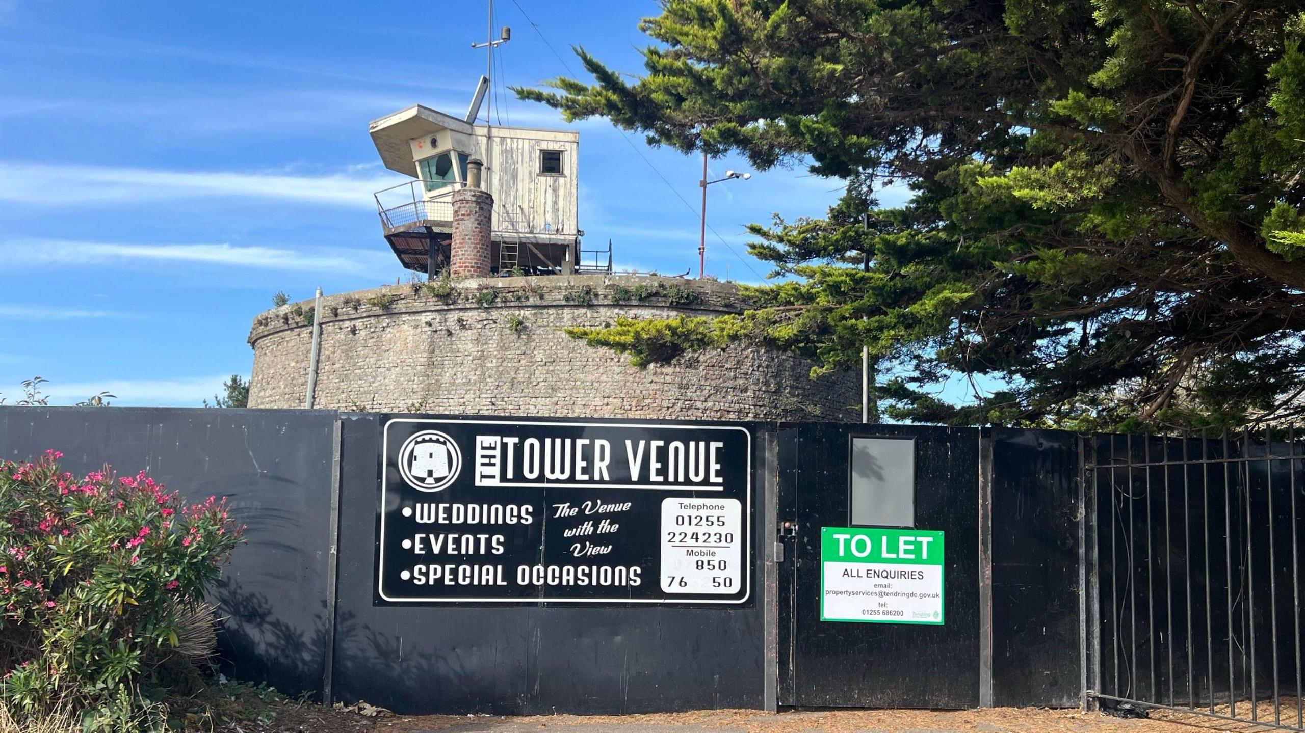 The circular brick fort is pictured behind black hoarding. It has a small wooden watchtower on top and a large tree to the right. On the black hoarding, which is preventing access, is a green "to let" sign and another black sign advertising it as a wedding venue.