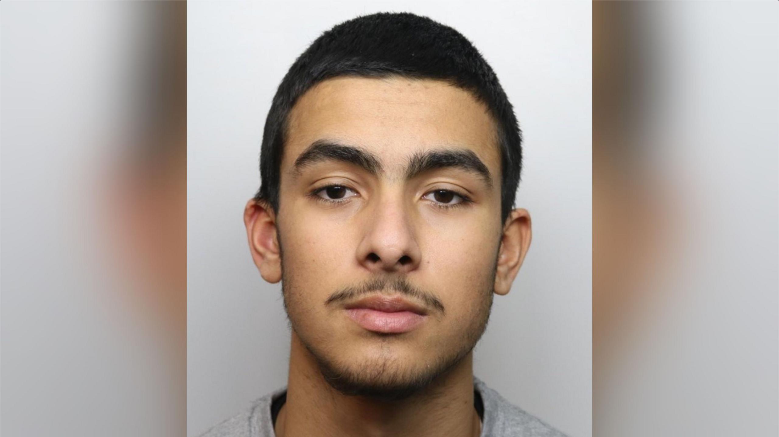 Tyler Wilkins with short dark hair and a slight beard wearing a grey top in a police custody photo