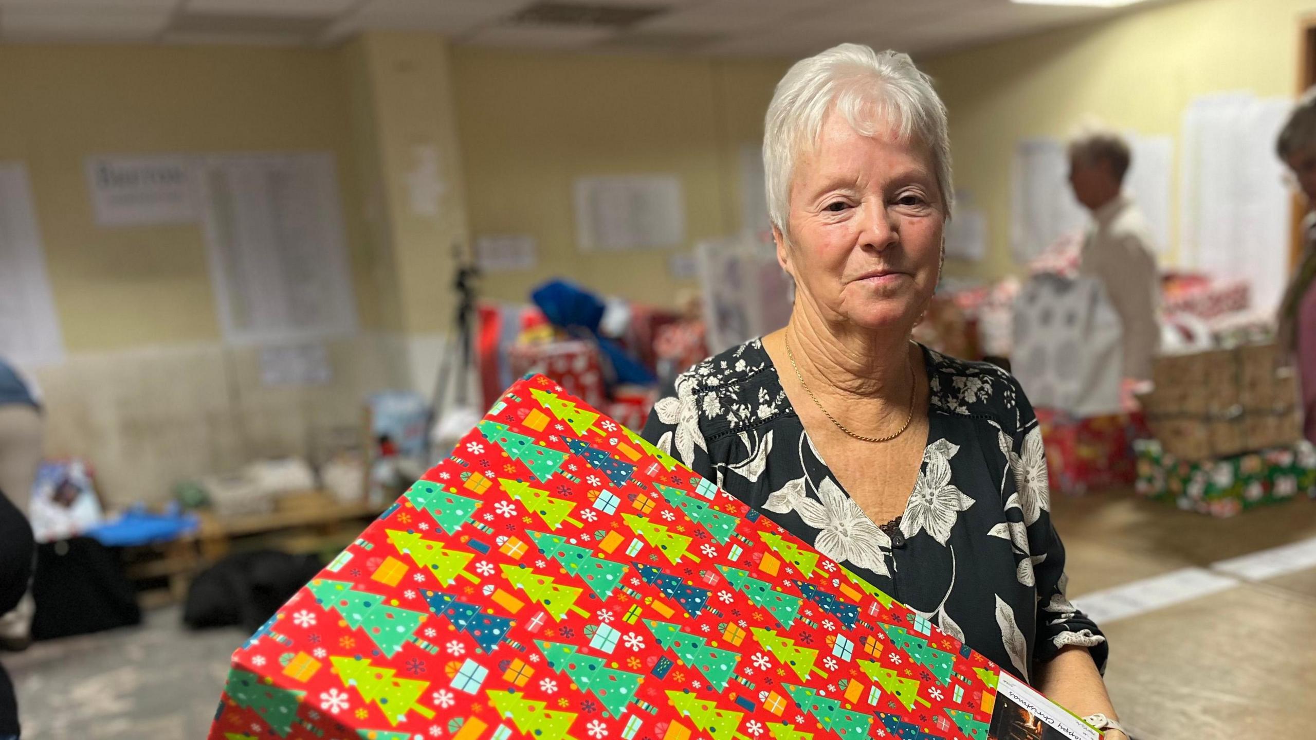 Jocelyn Dorey stands with a large present in front of her. She wears a black top with white flower on, while the present is wrapped in red paper with Christmas trees depicted in different shades of green