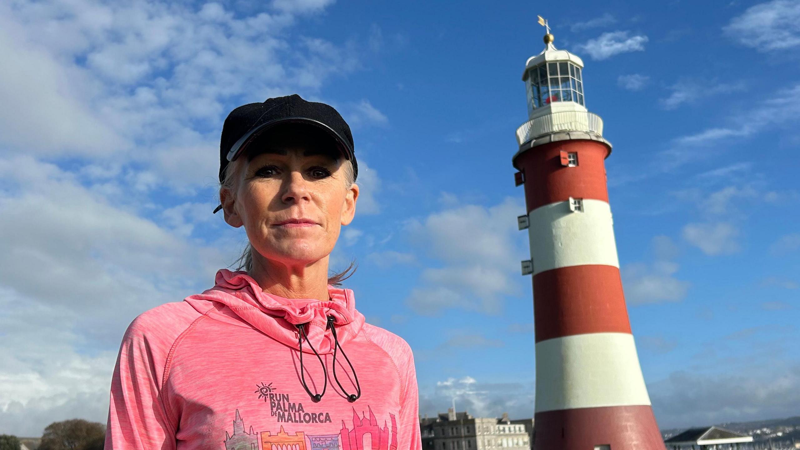 Nicky Putman is wearing a pink running jumper and a black cap on a bright sunny day in Plymouth. She is stood on grass looking straight at the camera. There is a lighthouse and the sea in the background. 