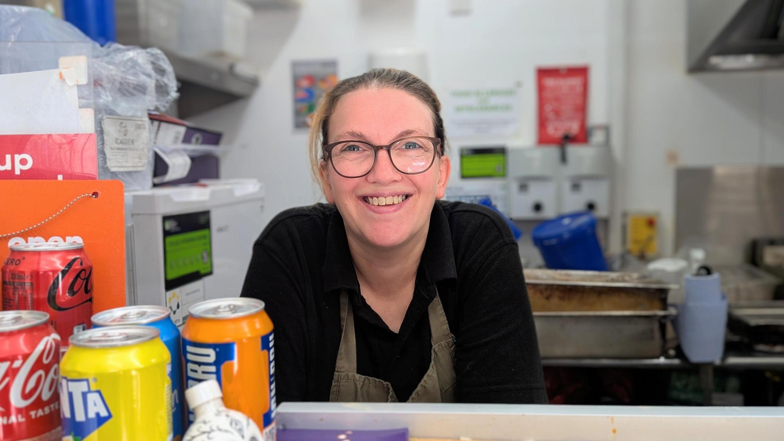 Rachel Stainton stands behind a counter in a food service area with drinks cans and other items on display. She is smiling, has blonde hair and wears glasses. Kitchen equipment and wall signs are visible in the background.