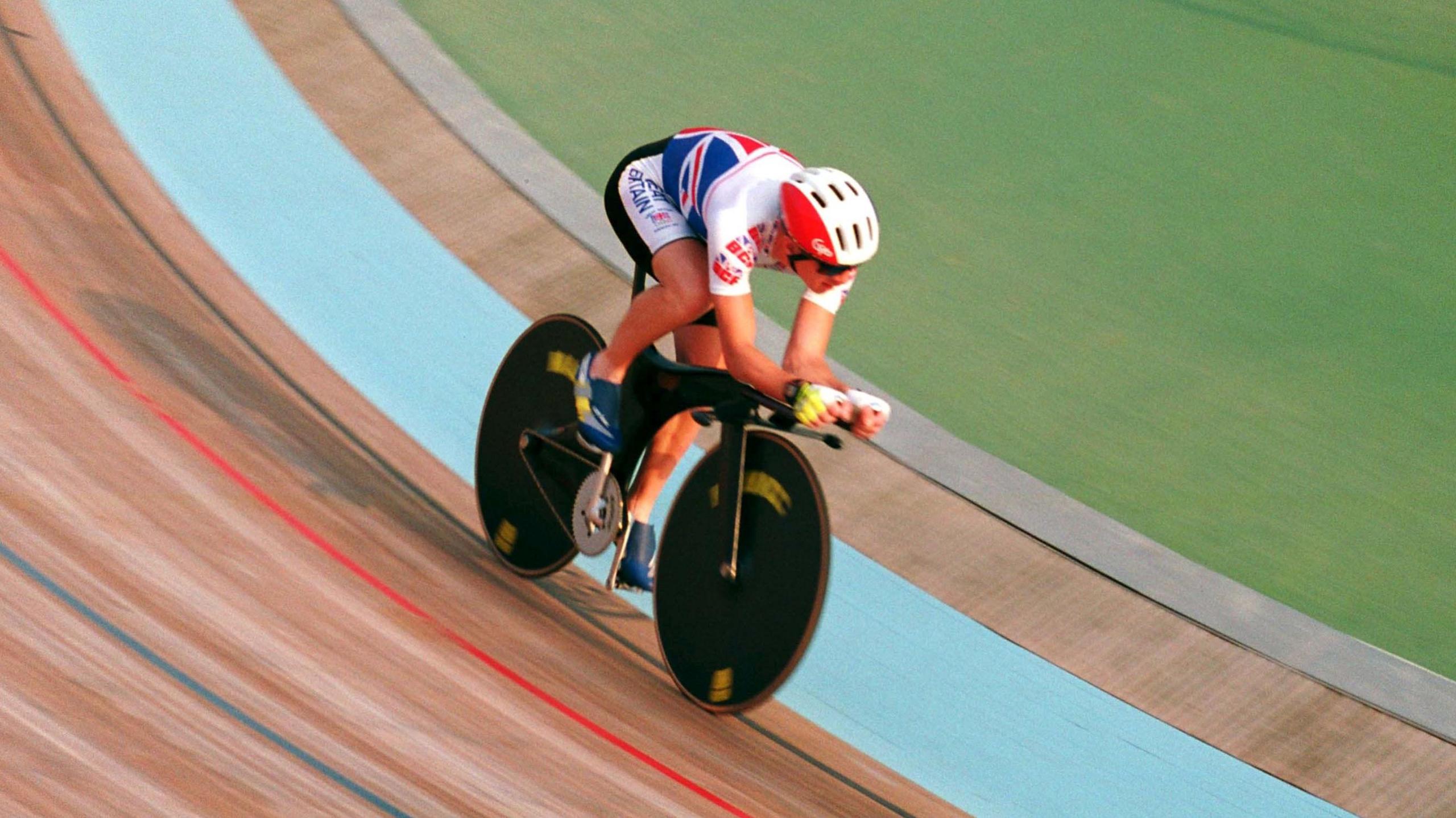 Chris Boardman on a bike racing around a velodrome circuit. He is wearing a red and white bike helmet and team GB's red, white, and blue colours on his top and bottom.