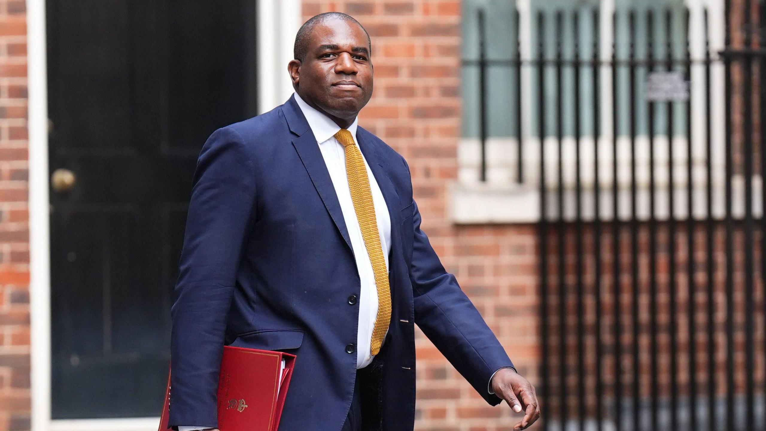 David Lammy, wearing a navy blazer, yellow tie and white shirt, walking and holding a red folder