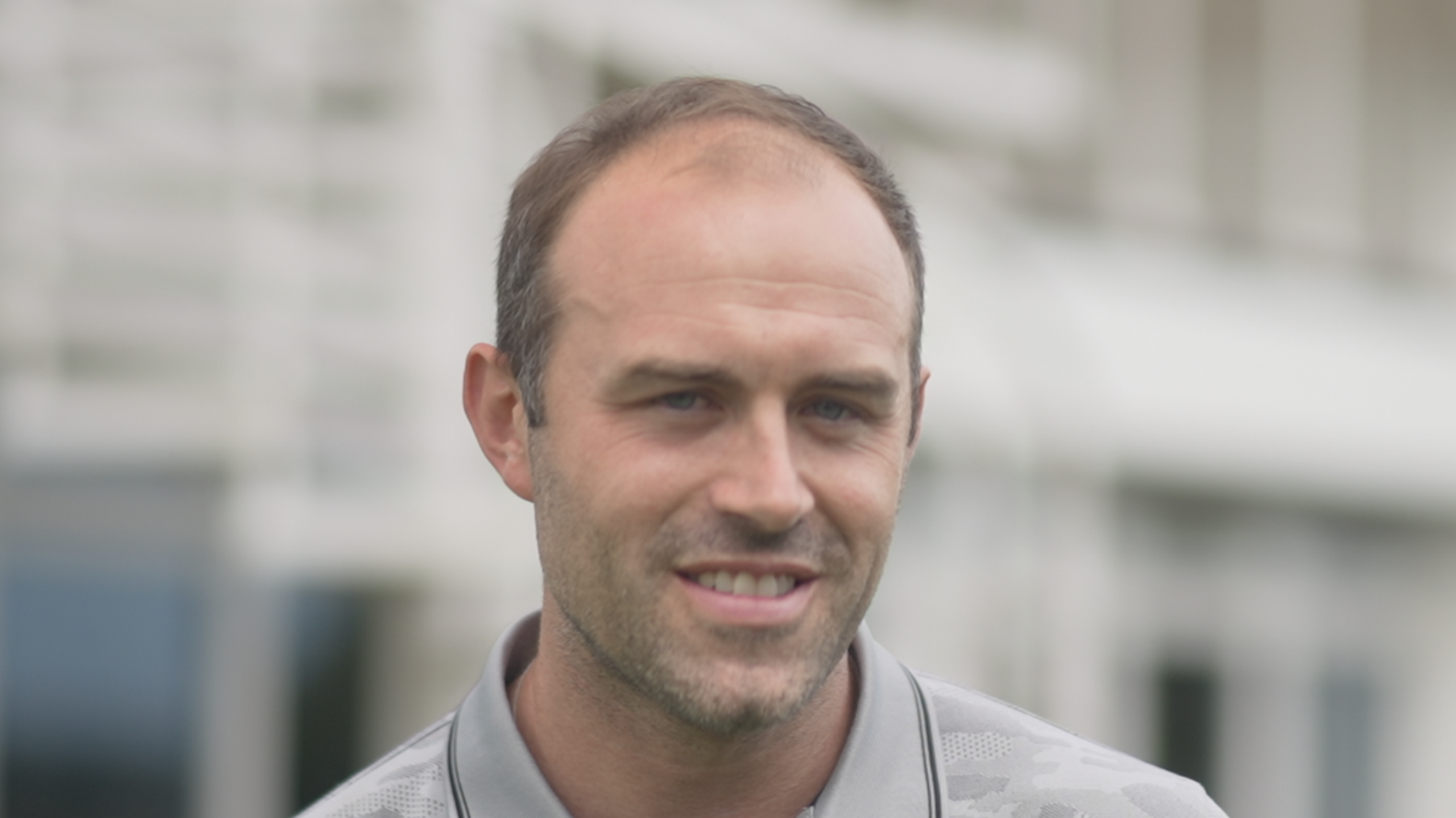 Jonny Sharkey smiles at the camera. He has thin dark hair that is receding. He wears a grey polo top.