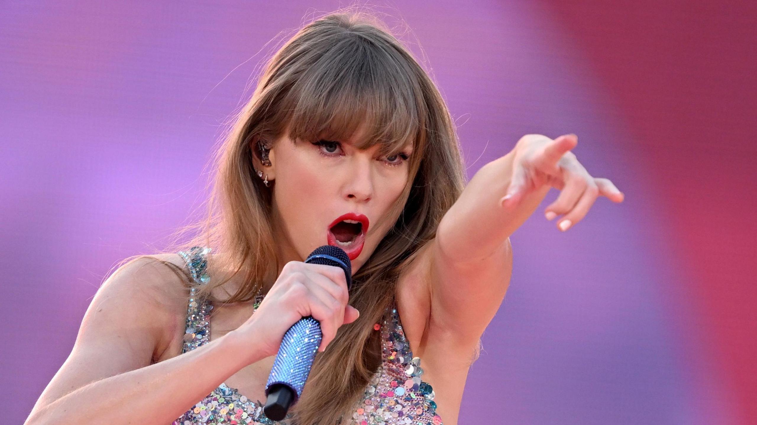 Taylor Swift performing on stage, singing into a microphone held up to her mouth with her right hand while pointing with her other hand at the audience. She has long straight hair, is wearing a sequin top and has a look of fierce determination on her face 