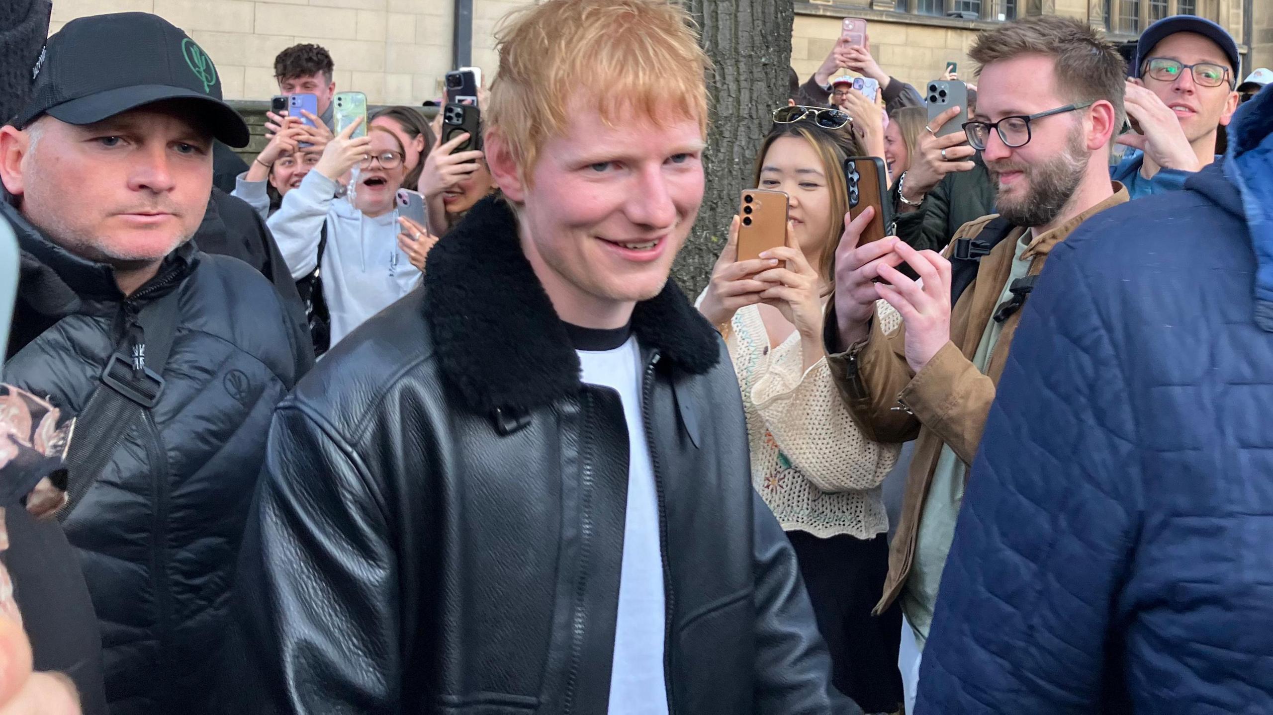 A ginger-haired man pushes through a crowd of people holding up phones. He is wearing a black leather jacket with fur around the collar and a white t-shirt.