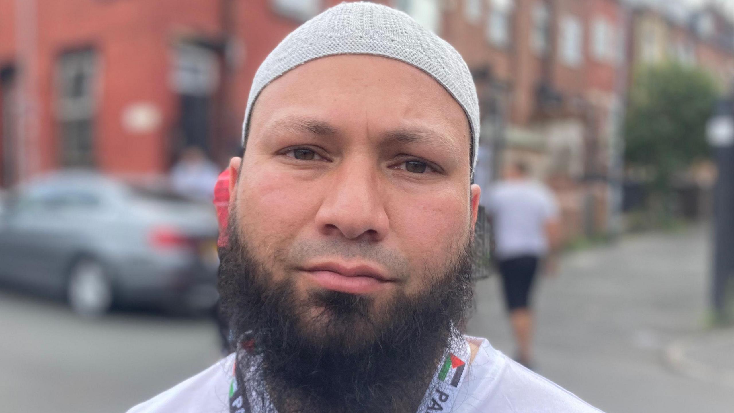 A man with a grey cap and black beard looks at the camera. He wears a white polo shirt and a lanyard reading "Palestine".