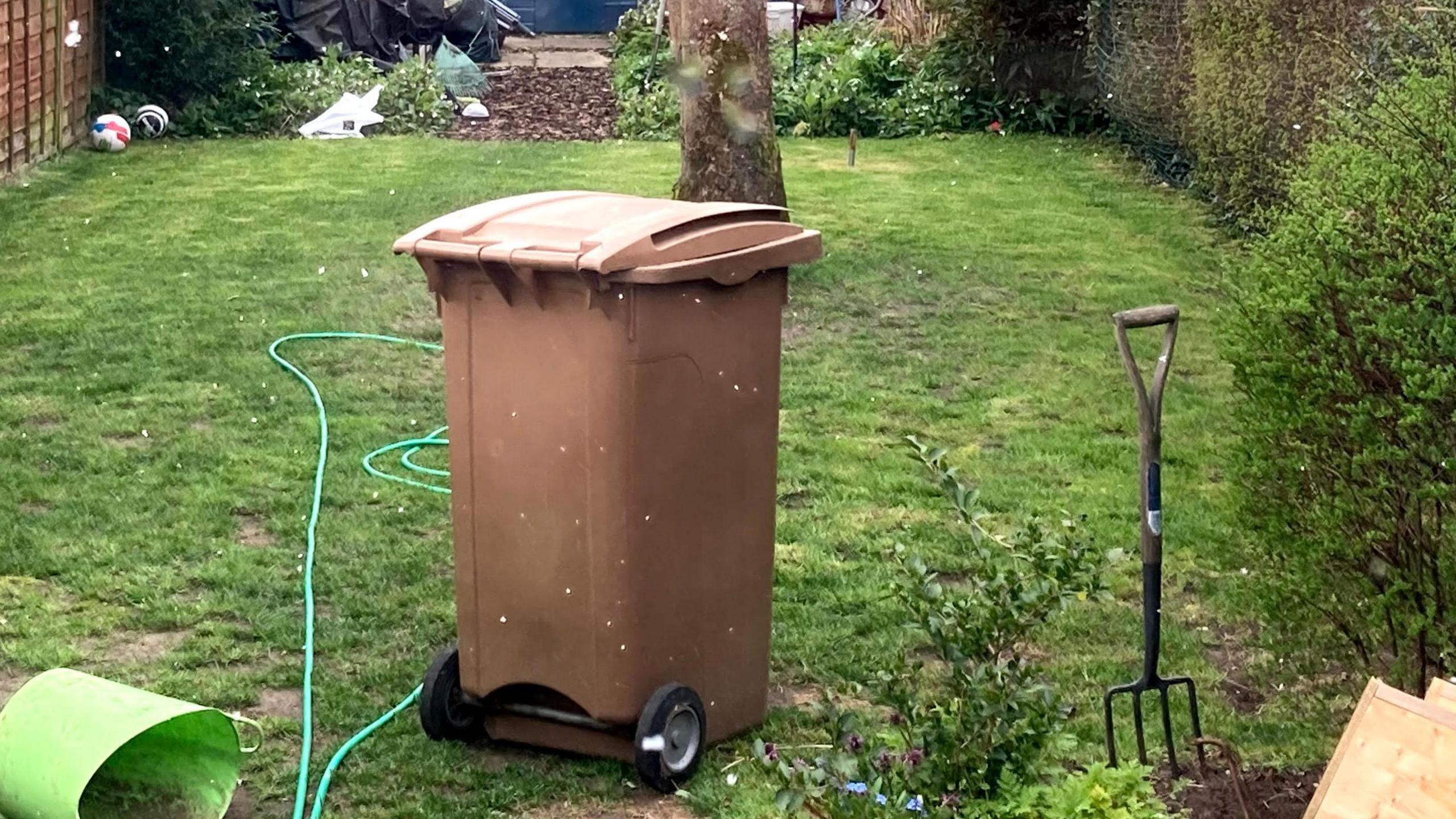 A brown garden waste bin sits in a garden. A green hose pipe lies on the grass around it as well as a garden fork.