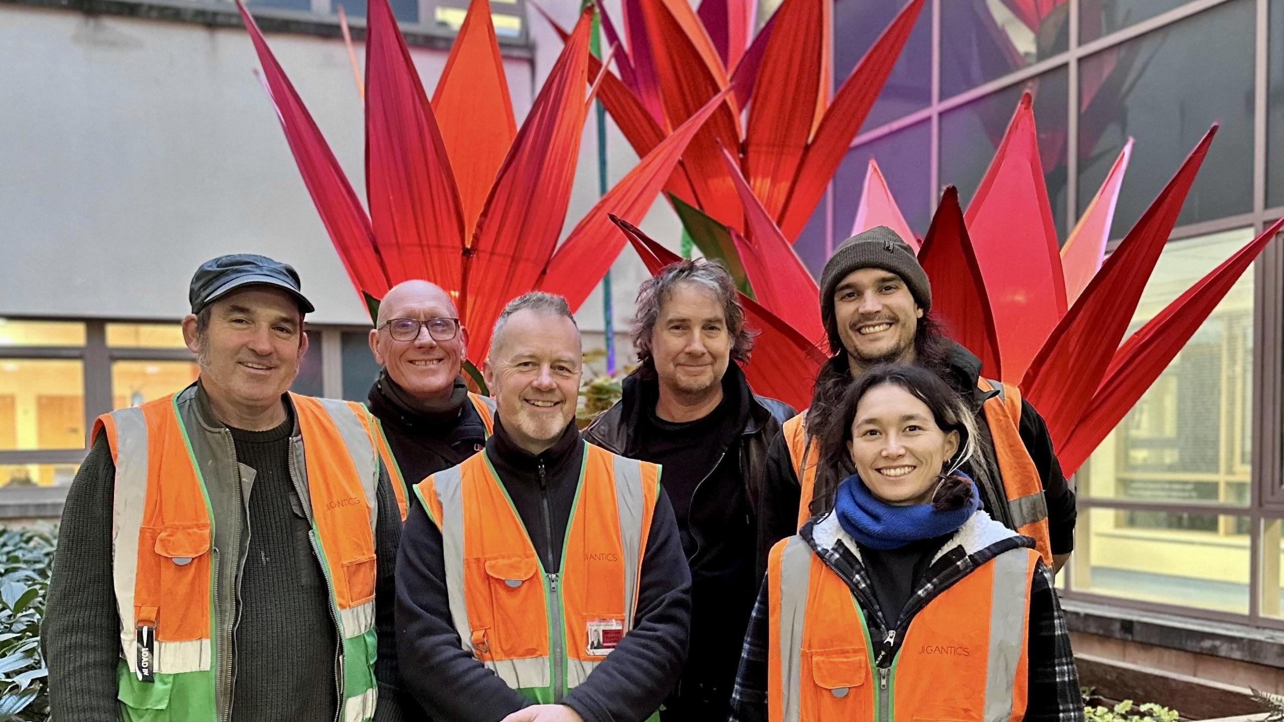 The team from Jigantics wearing orange hi-vis vests, standing in front of a large flower installation, which depicts red elongated petals growing upwards in a cone shape. The team of five men and one women are smiling at the camera.