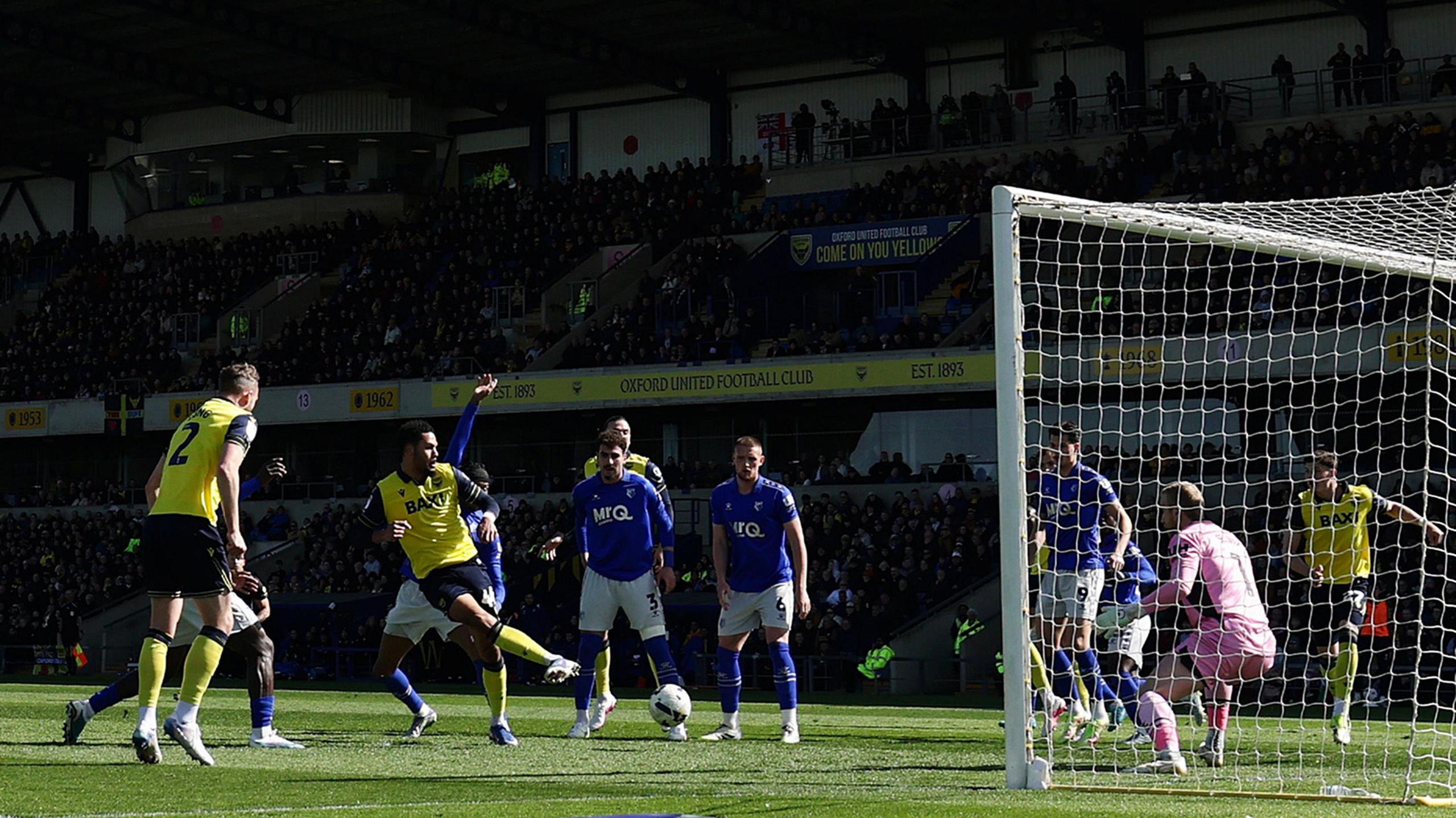 Footballer Myles Peart-Harris strikes a football for Oxford United in a crowded penalty area during a Championship match at home to Watford. The Oxford players are wearing a yellow kit with dark blue shorts and trim details, while Watford are wearing blue shirts and socks with white shorts. A goalkeeper in a predominantly pink kit is visible on the goal line, as is a packed stand full of fans at the Kassam Stadium