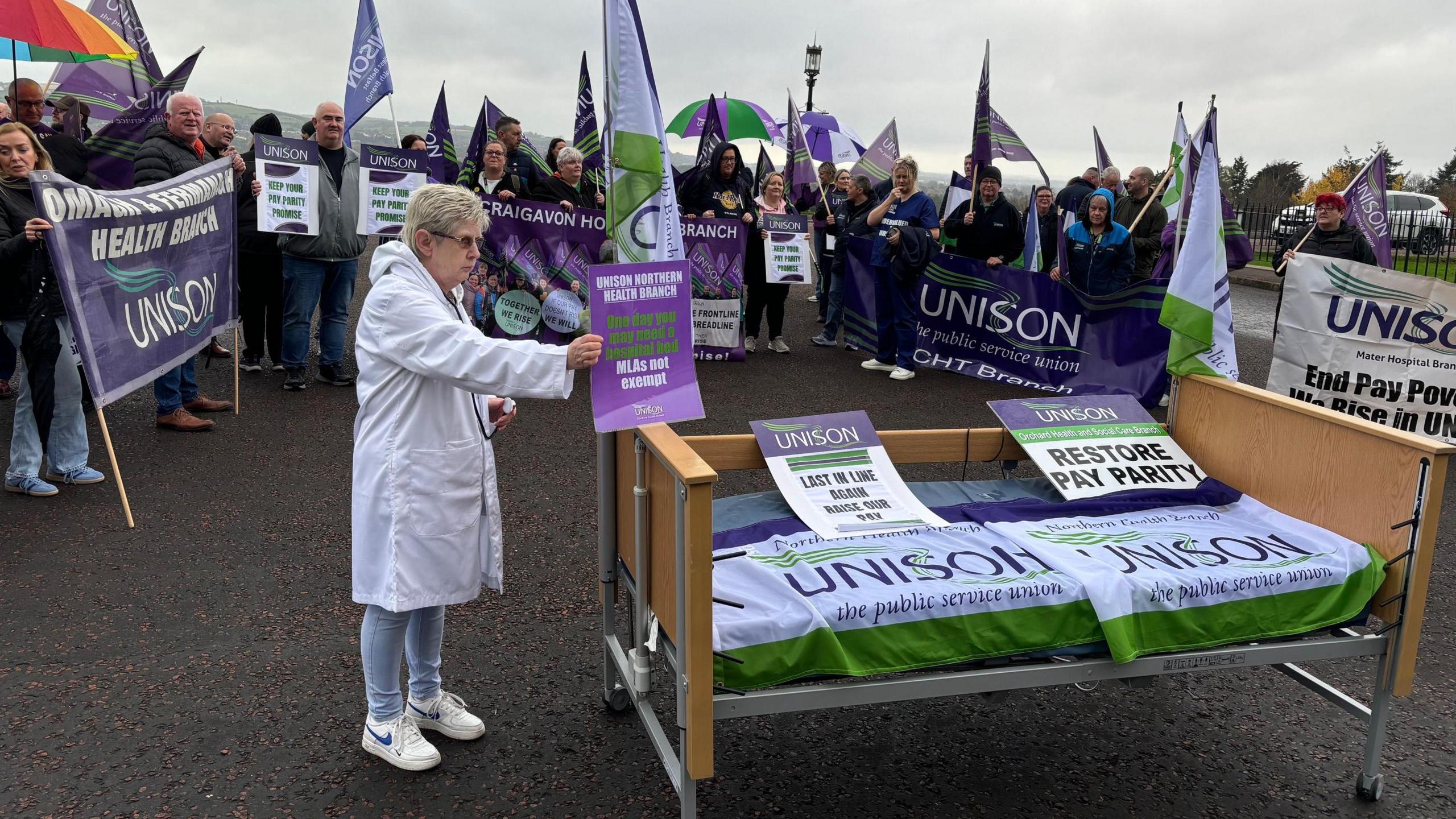 Multiple people holding purple and green unison flags and a hospital bed covered in unison signs sits outside Stormont and is being held on my a protestor. 