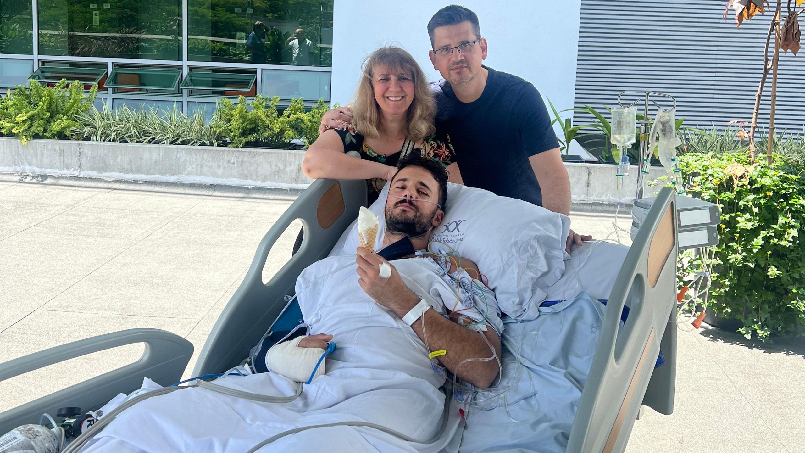 Luke Tarrant, lying in a hospital bed, outside, eating an ice cream, with a bandaged arm, tubes coming out his other arm, with his parents, Daniela and David Tarrant behind him and behind a bed. David has his arm around Daniela, he is wearing a dark T-shirt and Daniela, a floral dress, with long fair hair. David is wearing glasses. 